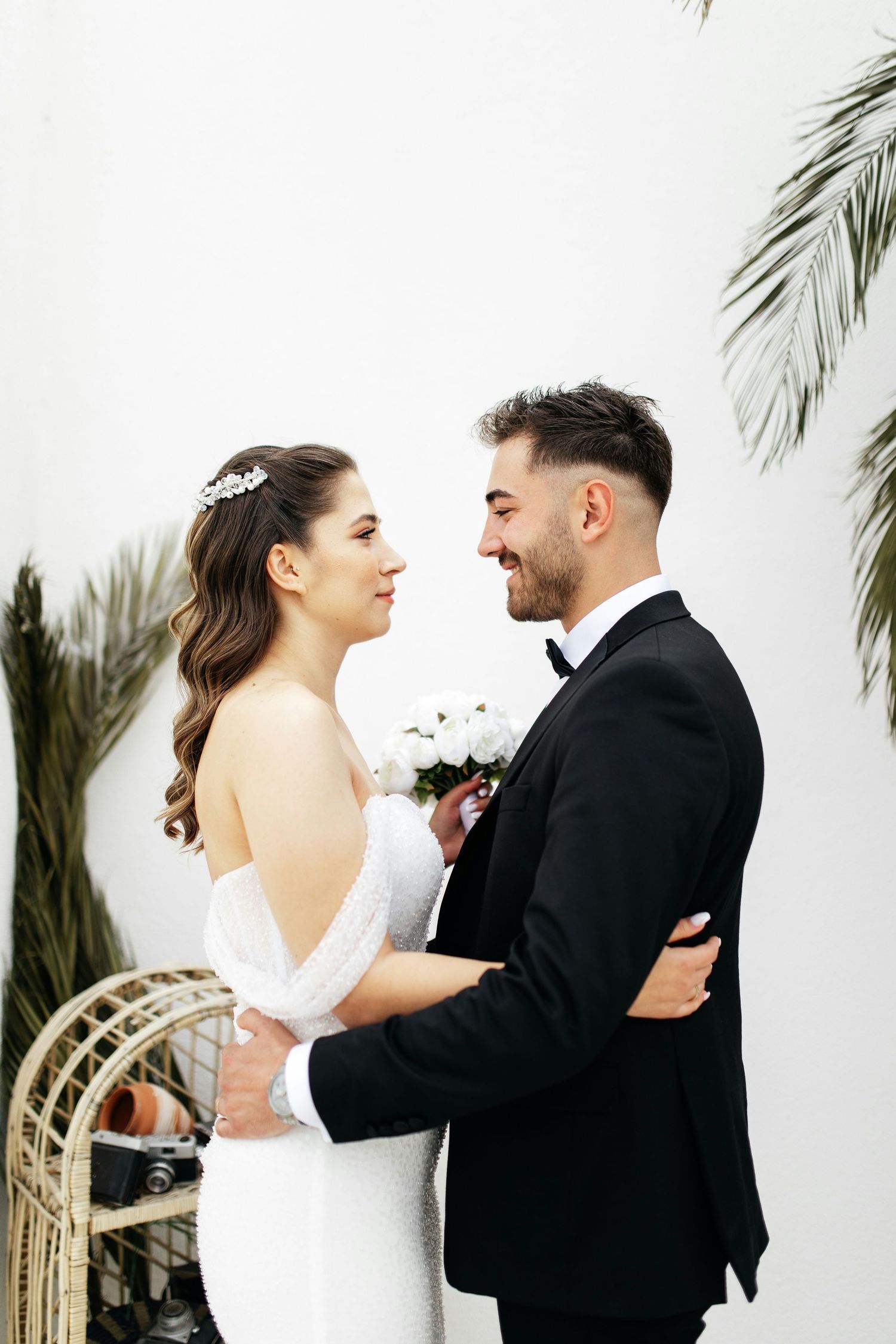 Smiling groom in black tuxedo embracing bride in white off-shoulder gown with tropical decor backdrop.