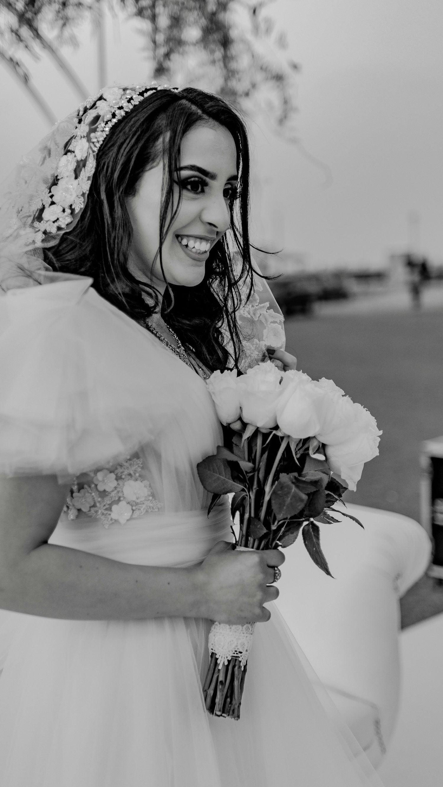 Black and white portrait of joyful bride holding rose bouquet wearing floral crown and veil.