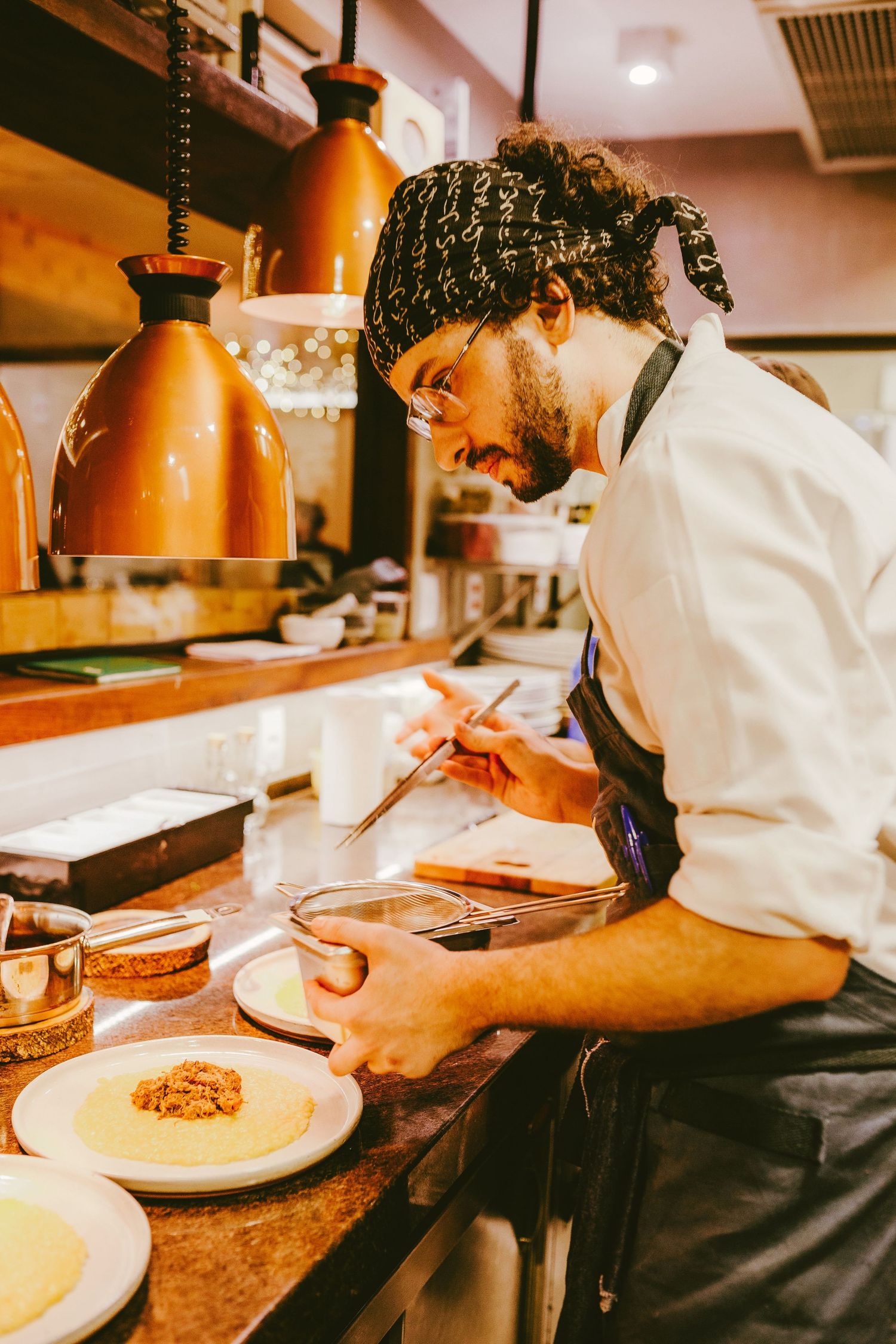 A chef in a bandana and apron carefully plates a dish in a warm, copper-lit restaurant kitchen.