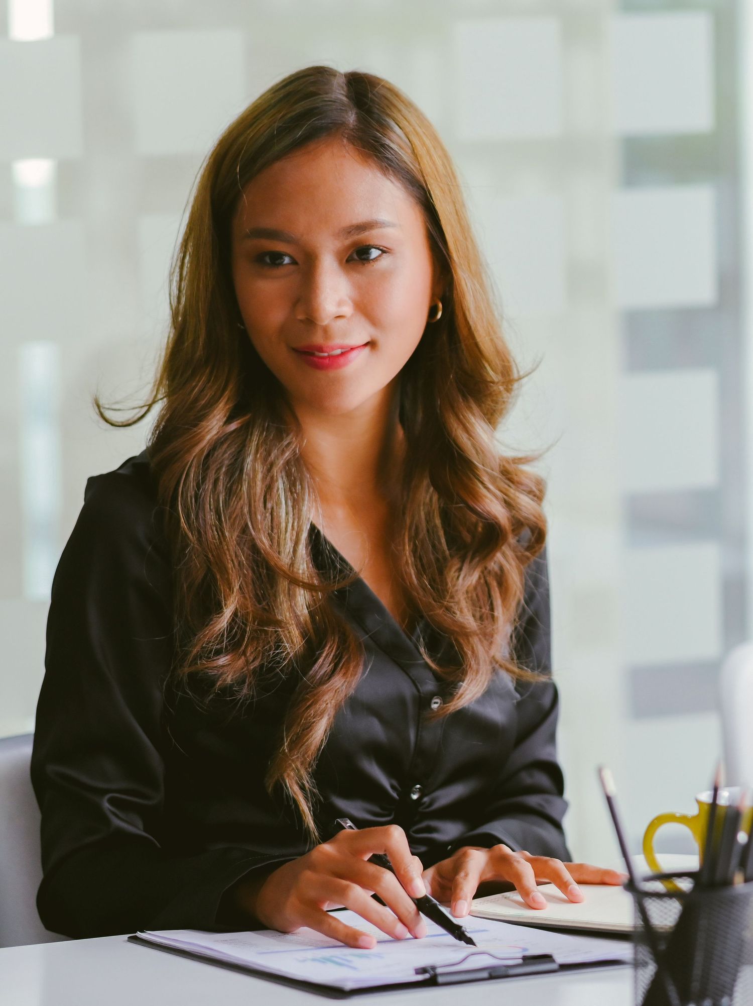 Professional businesswoman in black blazer signing documents at office desk, smiling confidently at camera.