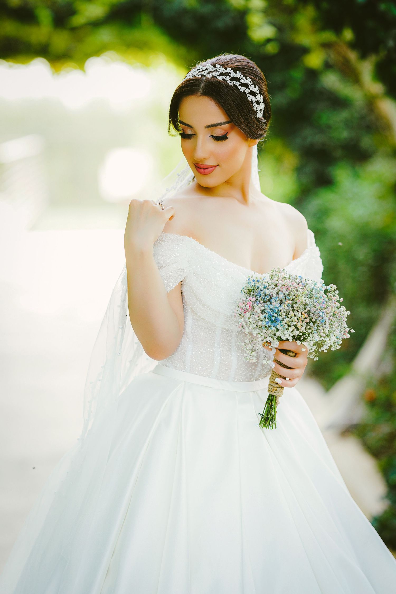 Elegant bride in white off-shoulder ball gown holding a pastel bouquet, wearing a floral headpiece outdoors.