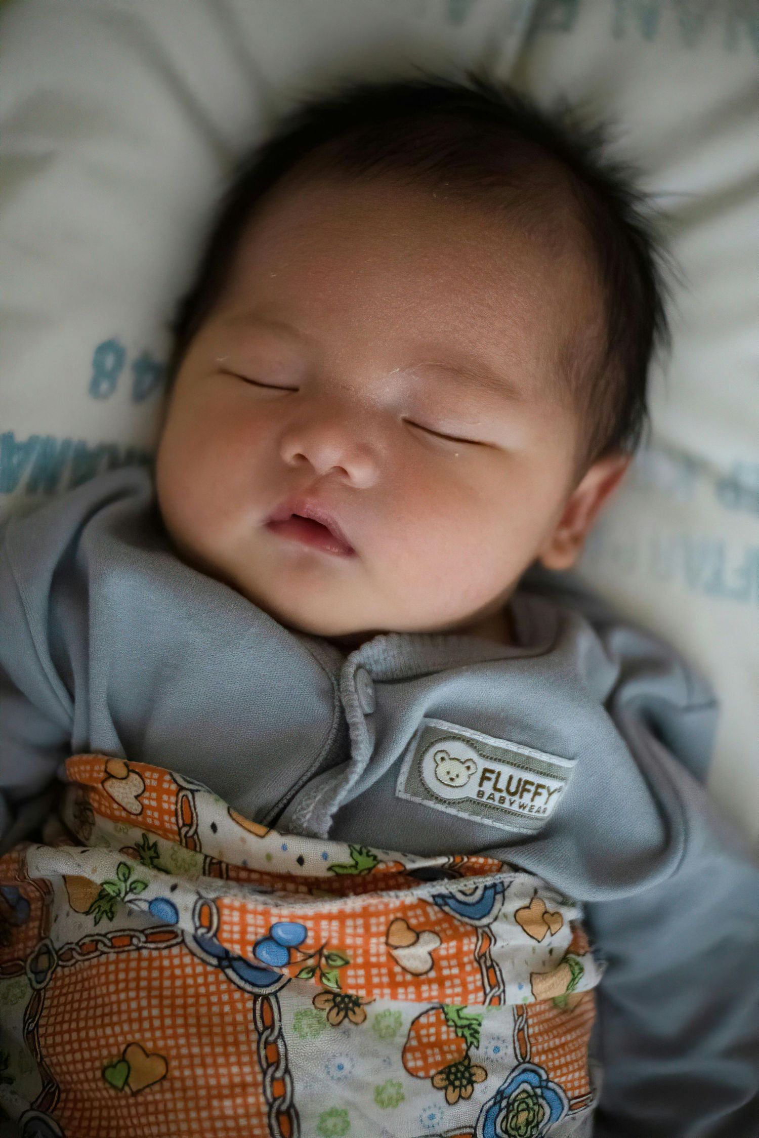 Sleeping Asian newborn baby swaddled in a gray Fluffy brand wrap on a soft blanket.