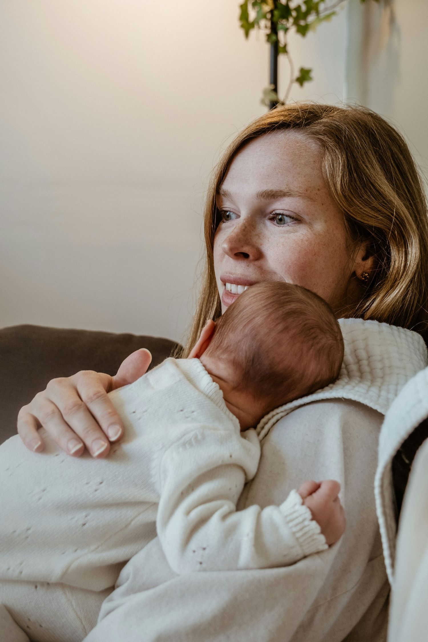Smiling mother with freckles tenderly holding sleeping newborn baby in white knit outfit.