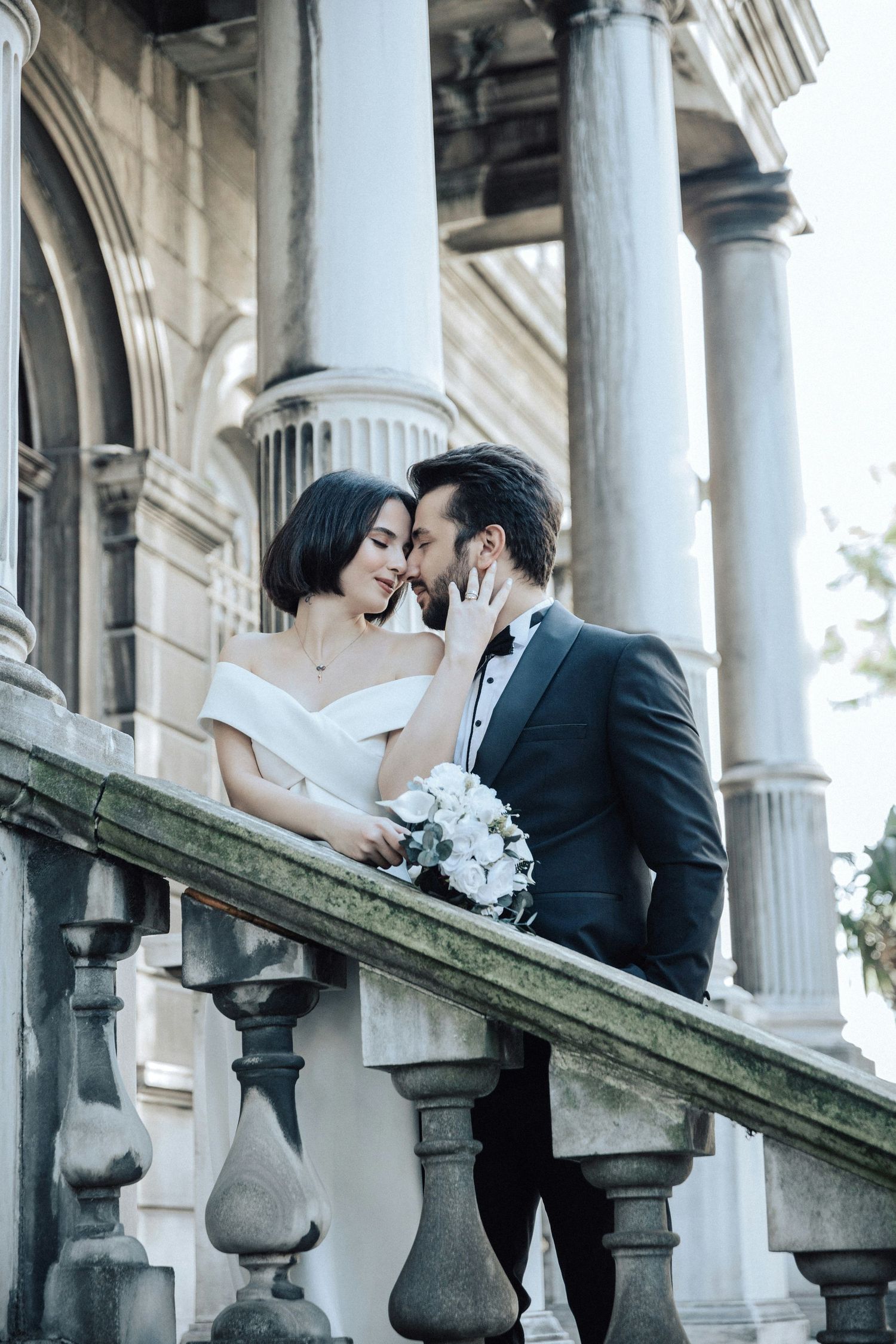 Bride and groom nearly kiss on ornate stone balcony of classical building, bride holding white floral bouquet.