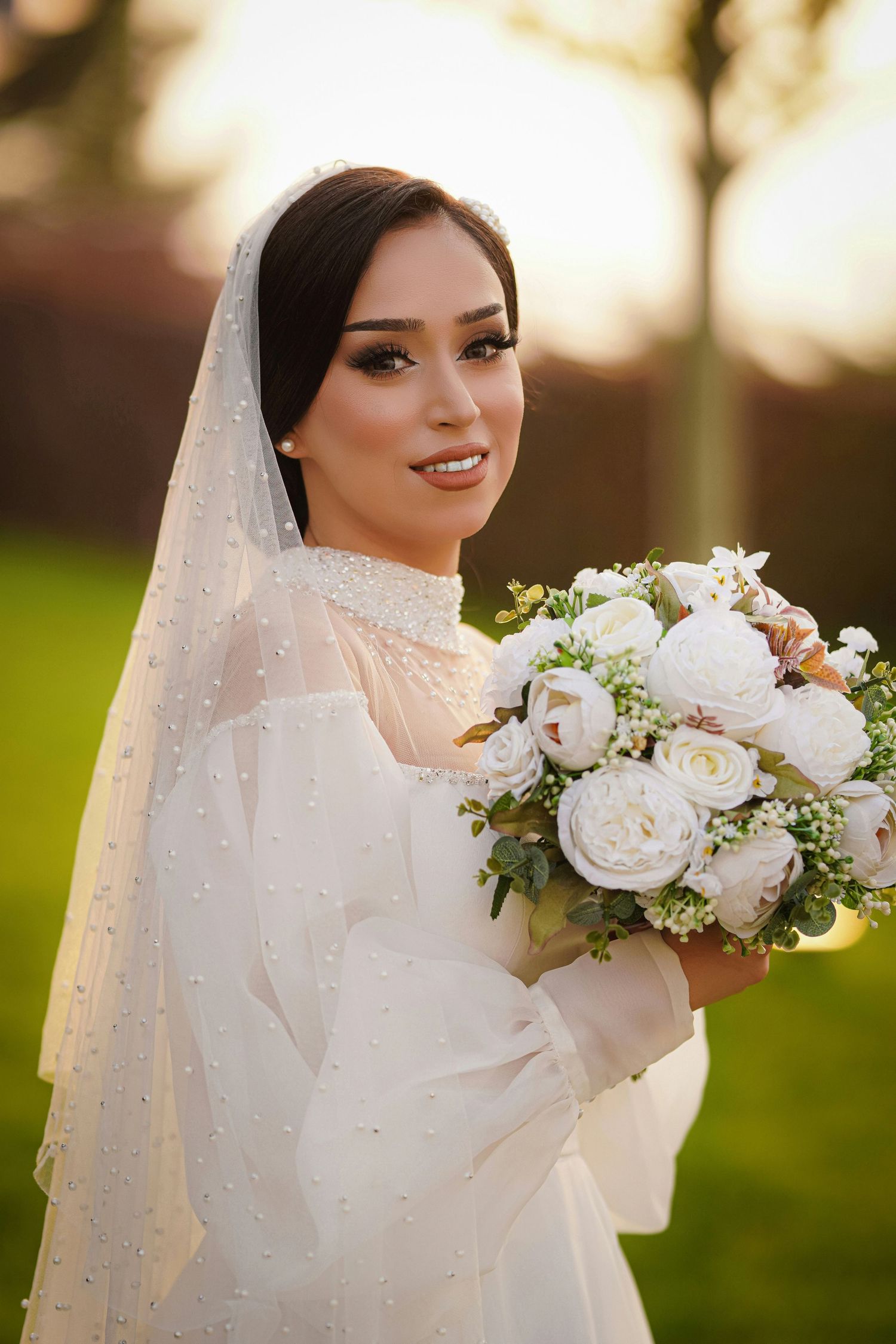 Elegant bride wearing pearl-embellished veil holding white peony bouquet in golden sunset light.