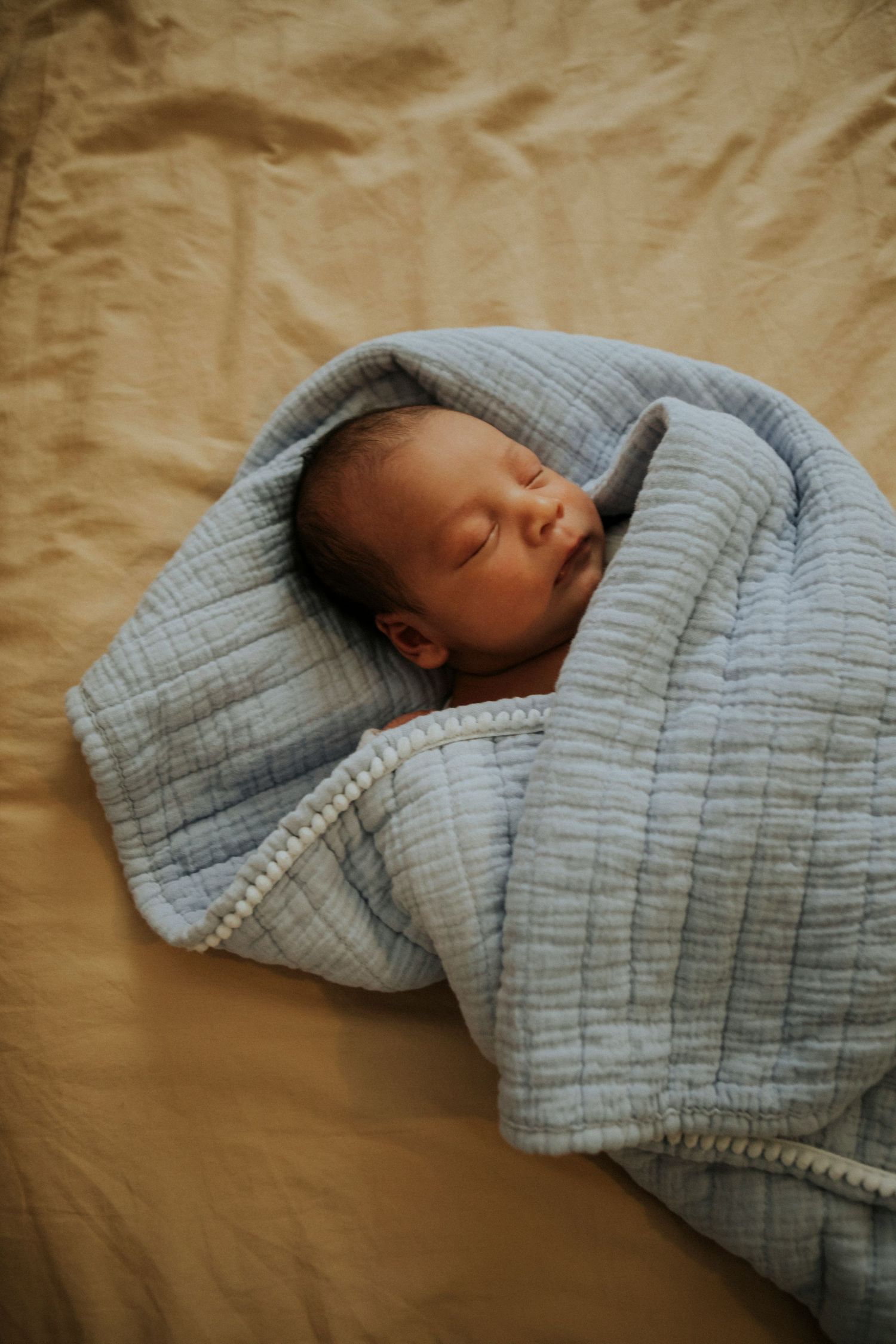 Newborn baby sleeping peacefully wrapped in a soft blue muslin blanket on a beige linen surface.