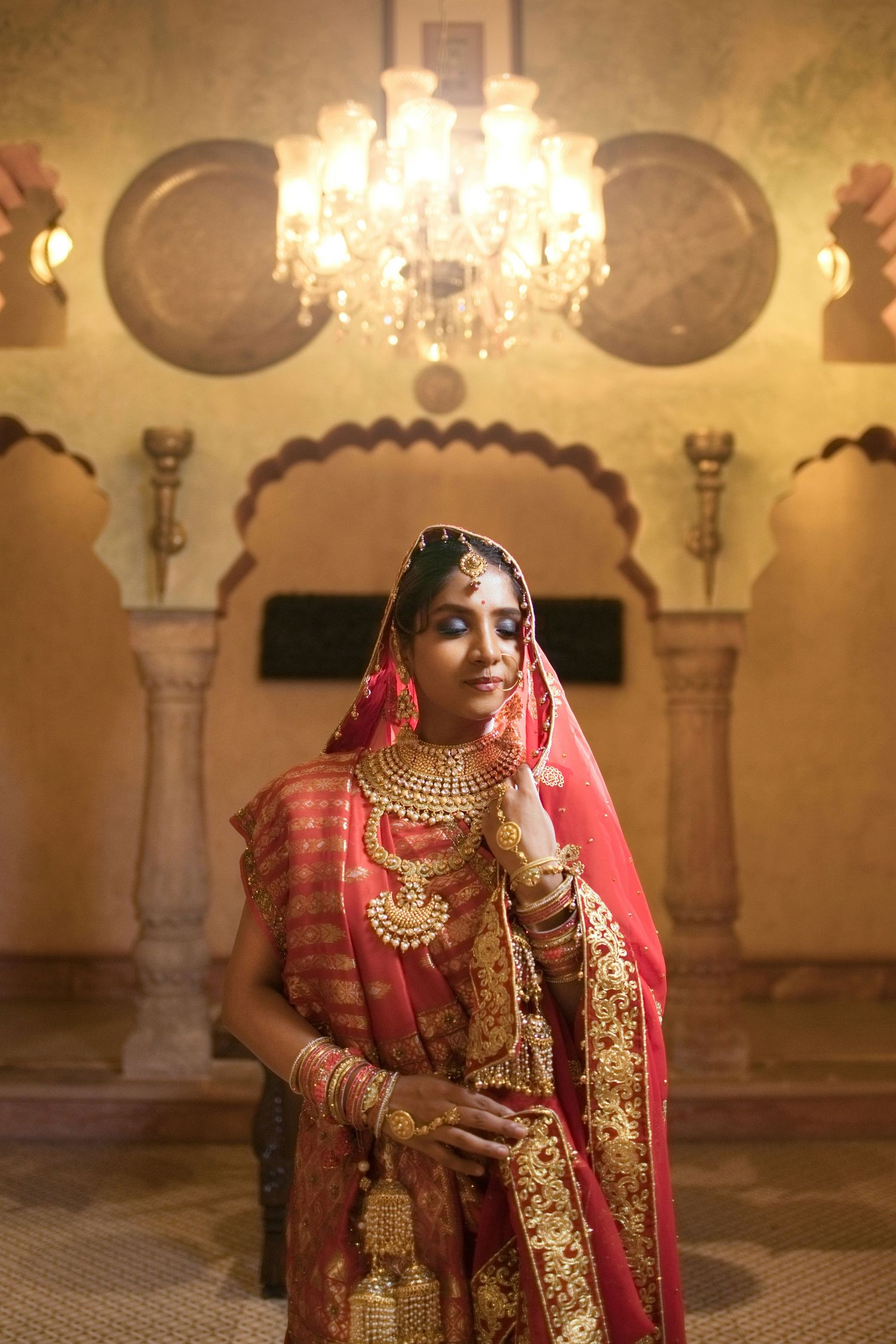 Indian bride in red and gold bridal lehenga with jewelry standing in ornate palace interior.