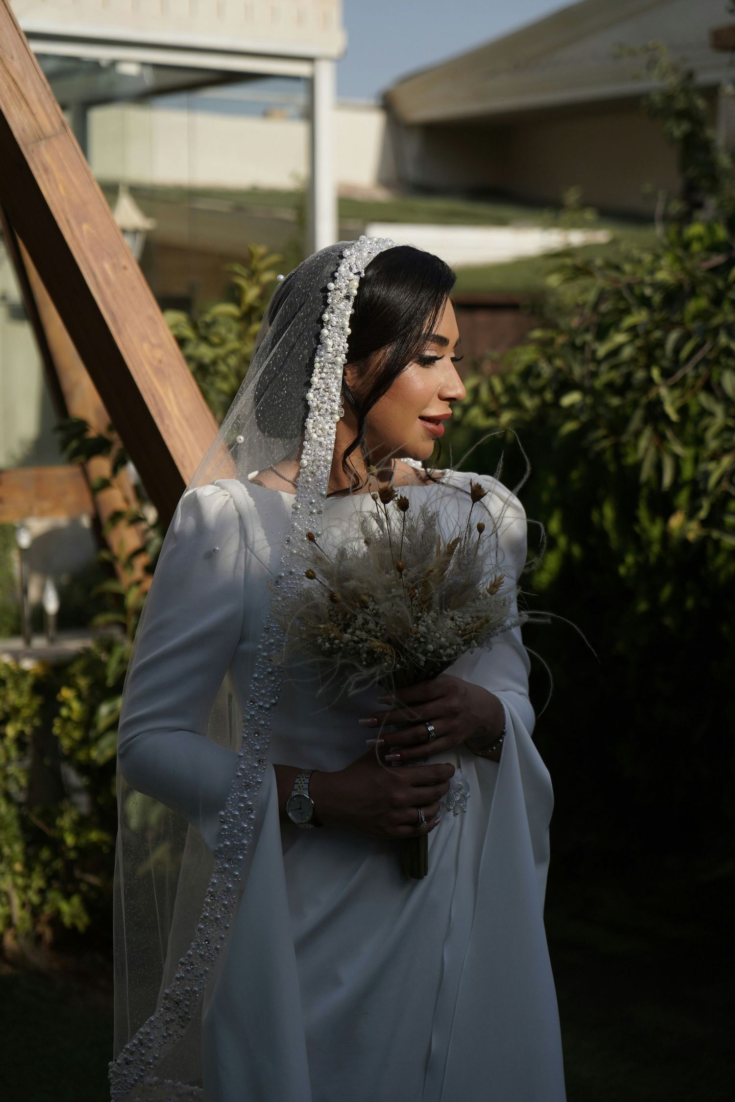 Elegant bride in white satin gown with embroidered veil holding a dried floral bouquet.