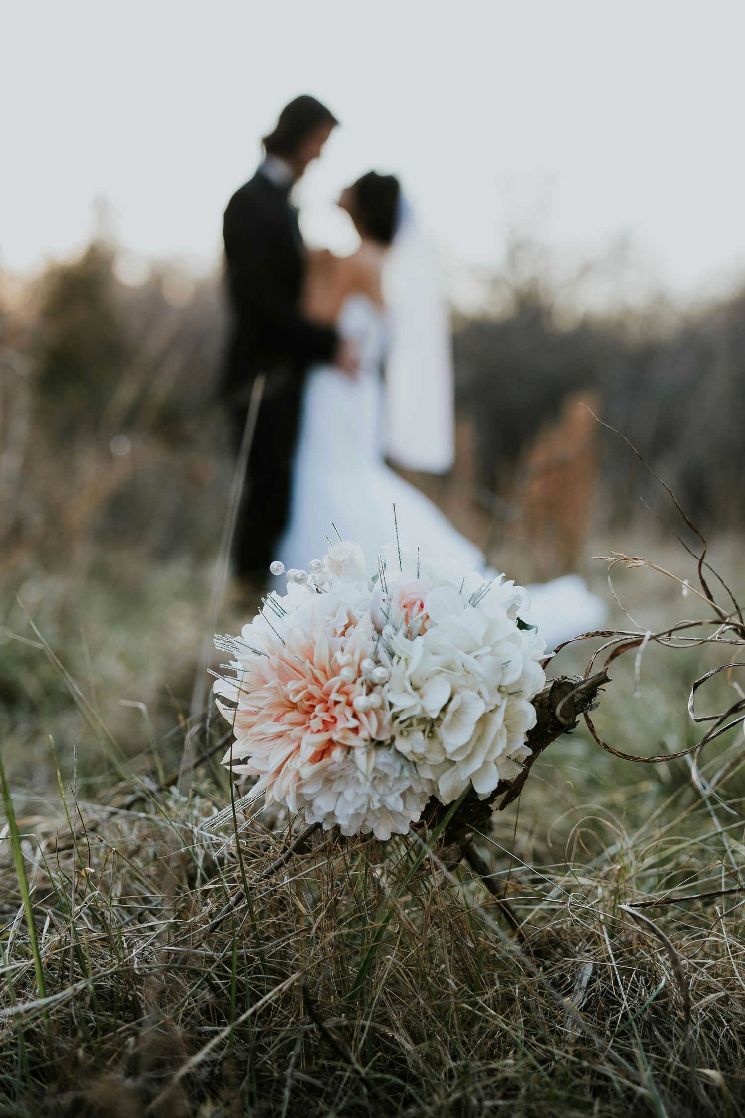 Wedding bouquet of pink dahlias and white flowers in grass with blurred bride and groom embracing behind.