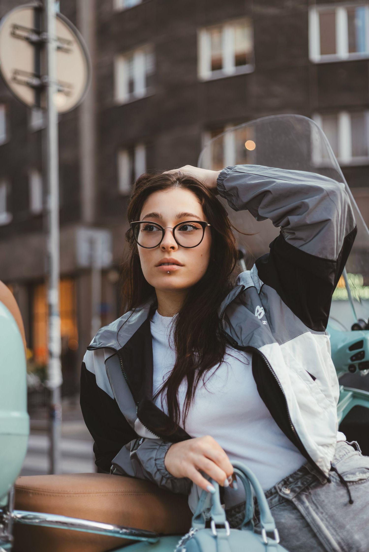 Young woman with glasses in urban jacket posing near scooter on city street.