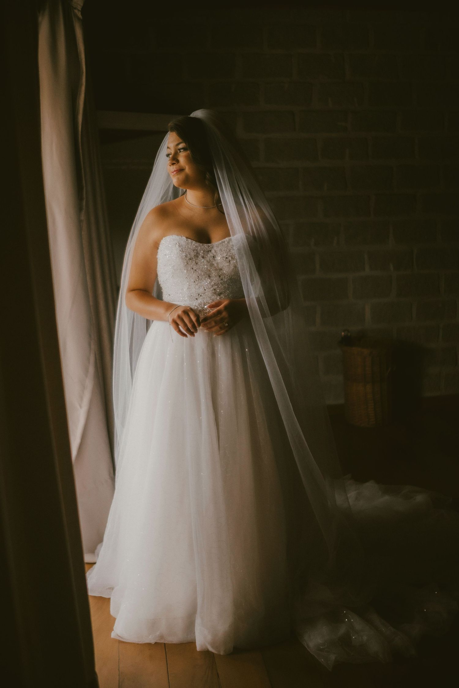Bride in strapless lace ballgown with long cathedral veil stands by window in moody light against brick wall.