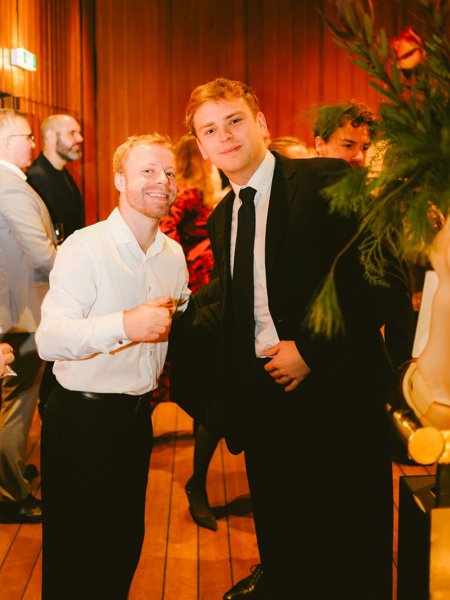 Two men in formal attire smile at a festive indoor event with warm lighting and guests in the background.