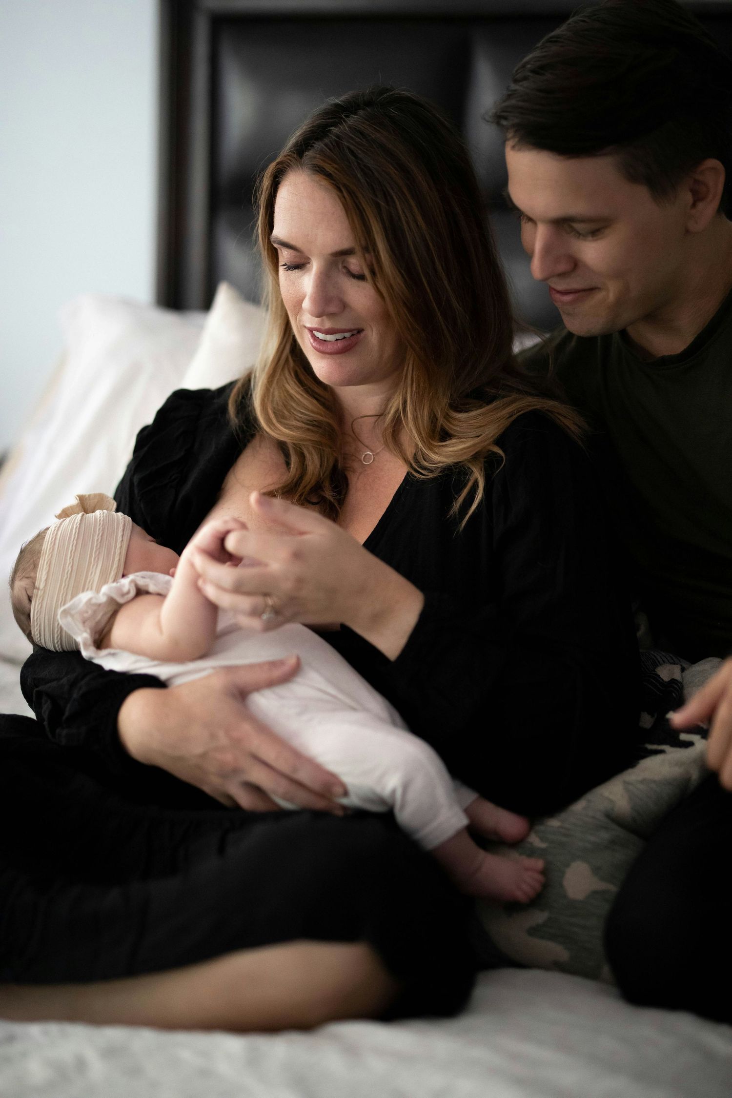A couple sits on a bed as the mother in black holds their newborn while the father looks on lovingly.