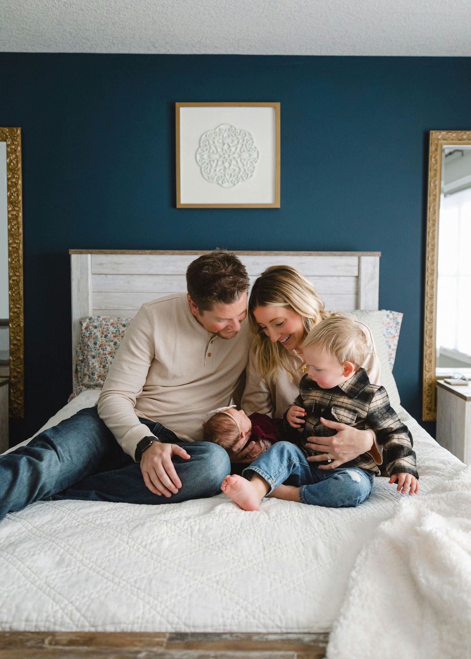A family of four sits on a bed with a navy blue wall, parents and toddler admiring their newborn.