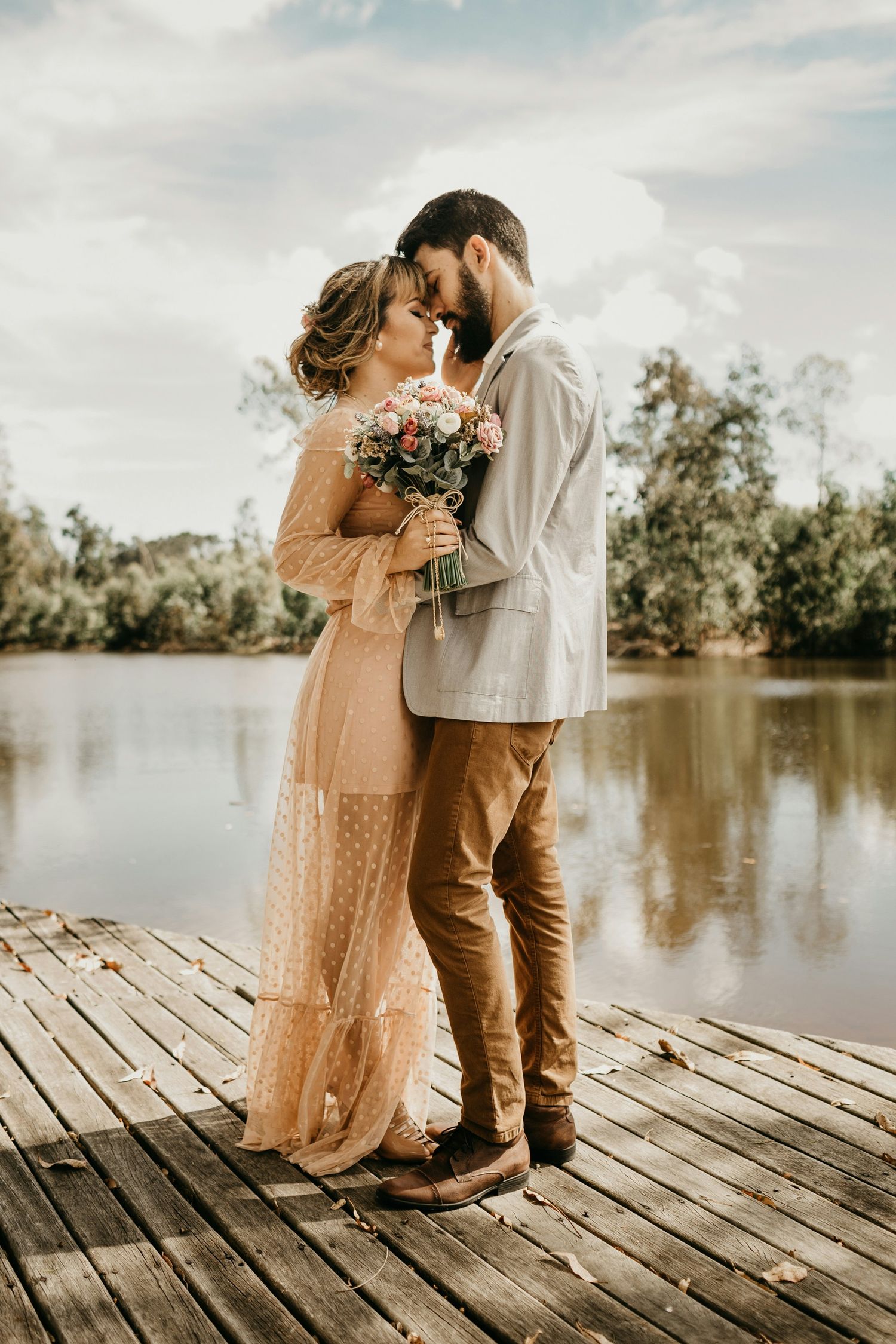 Bride in peach lace gown and groom in cream jacket touch foreheads on a wooden dock overlooking a serene lake.