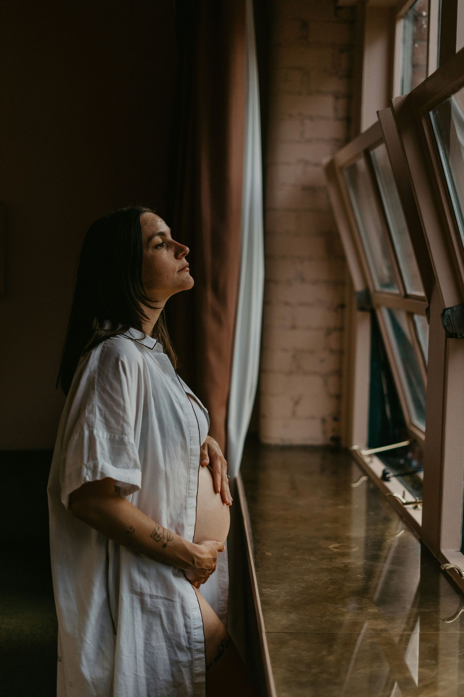Pregnant woman in white linen shirt standing by tall windows with eyes closed peacefully.