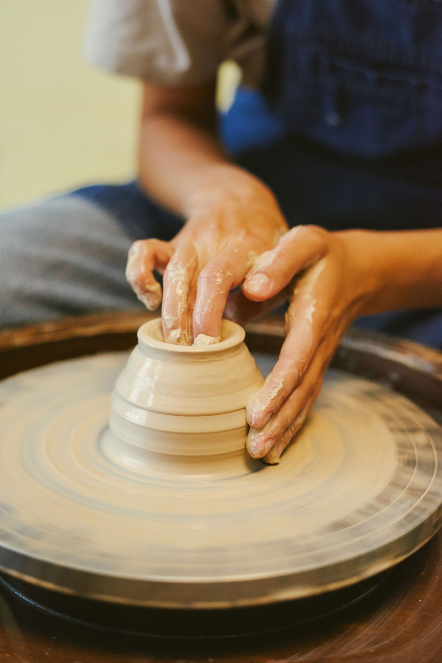 Hands shaping wet clay on a spinning pottery wheel, forming a small bowl in a ceramics studio.
