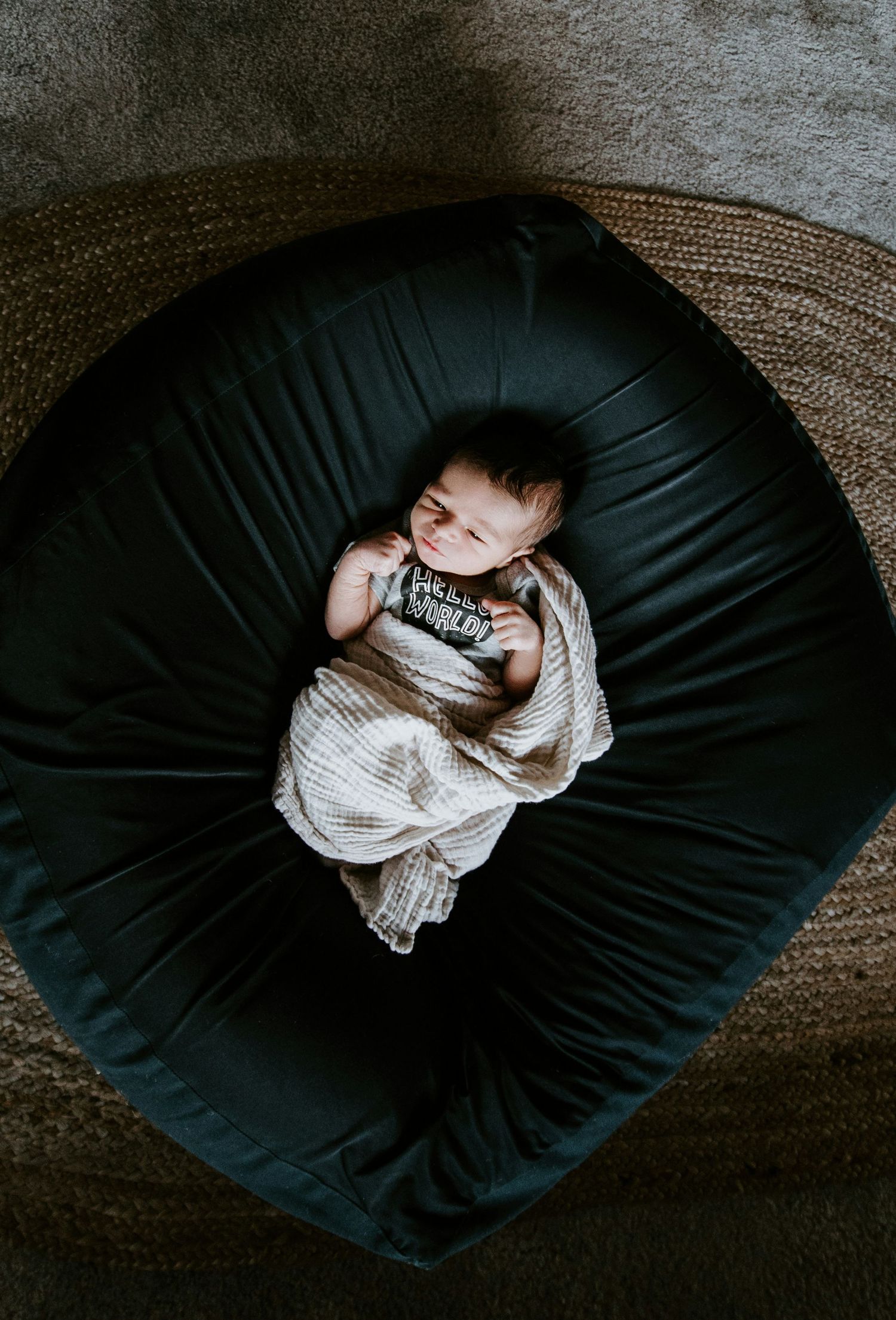 Sleeping newborn baby wrapped in muslin swaddle on dark bean bag chair, top view.