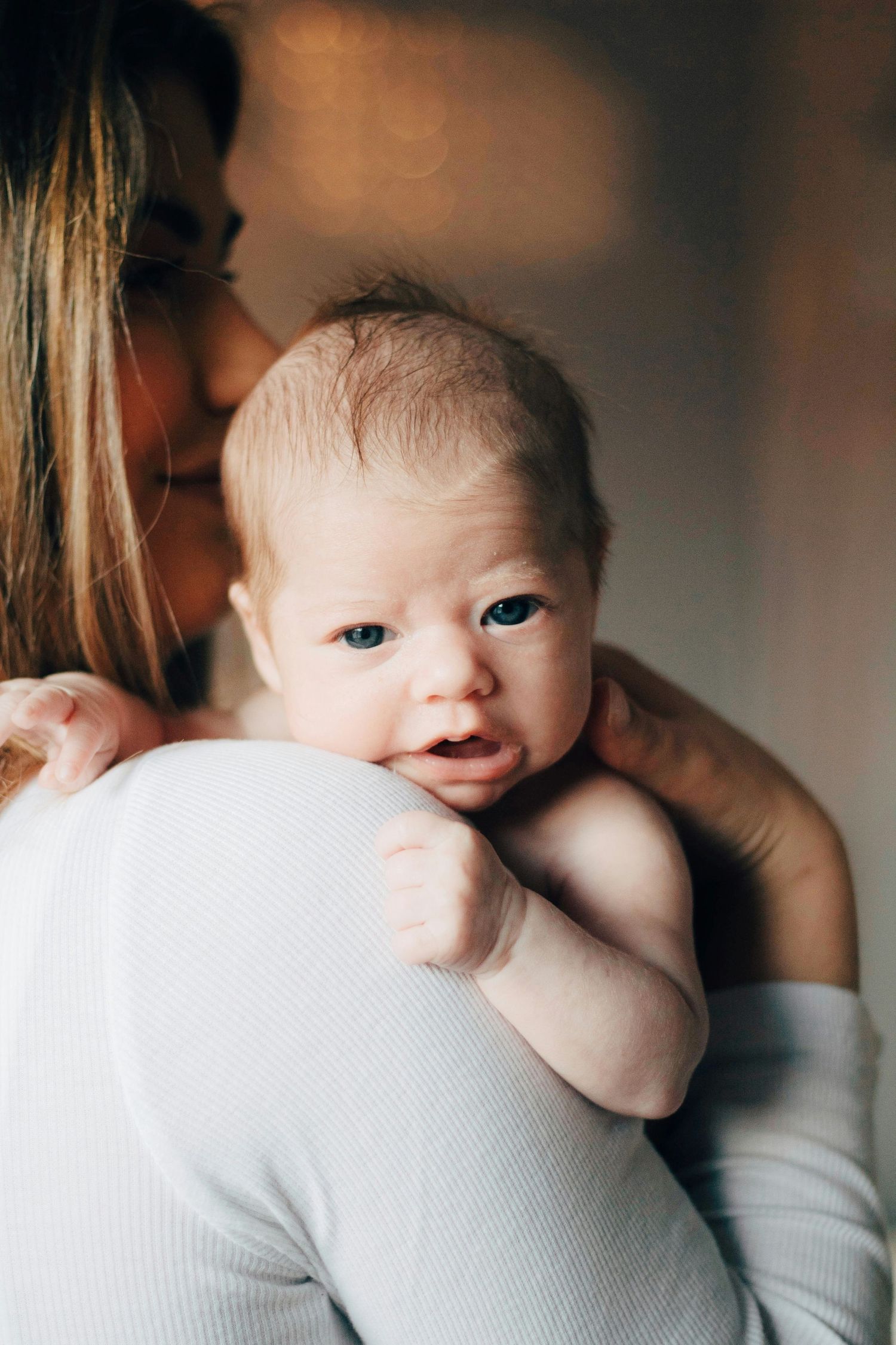 A mother tenderly holds her newborn baby against her shoulder, the infant gazing curiously at the camera.