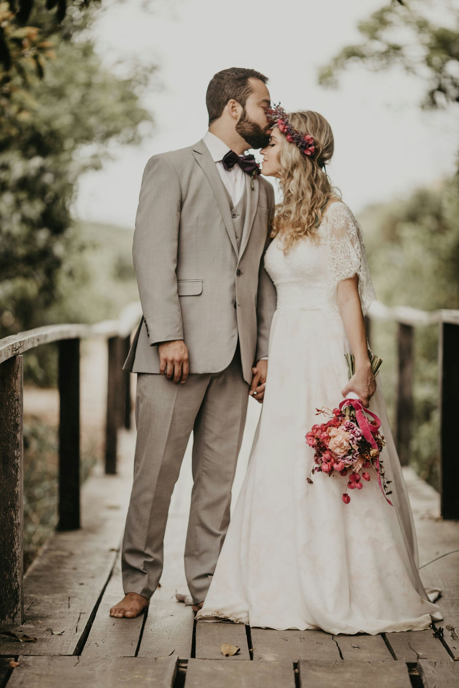 Bride and groom kiss on a wooden bridge, bride wearing a floral crown and holding a vibrant pink bouquet.
