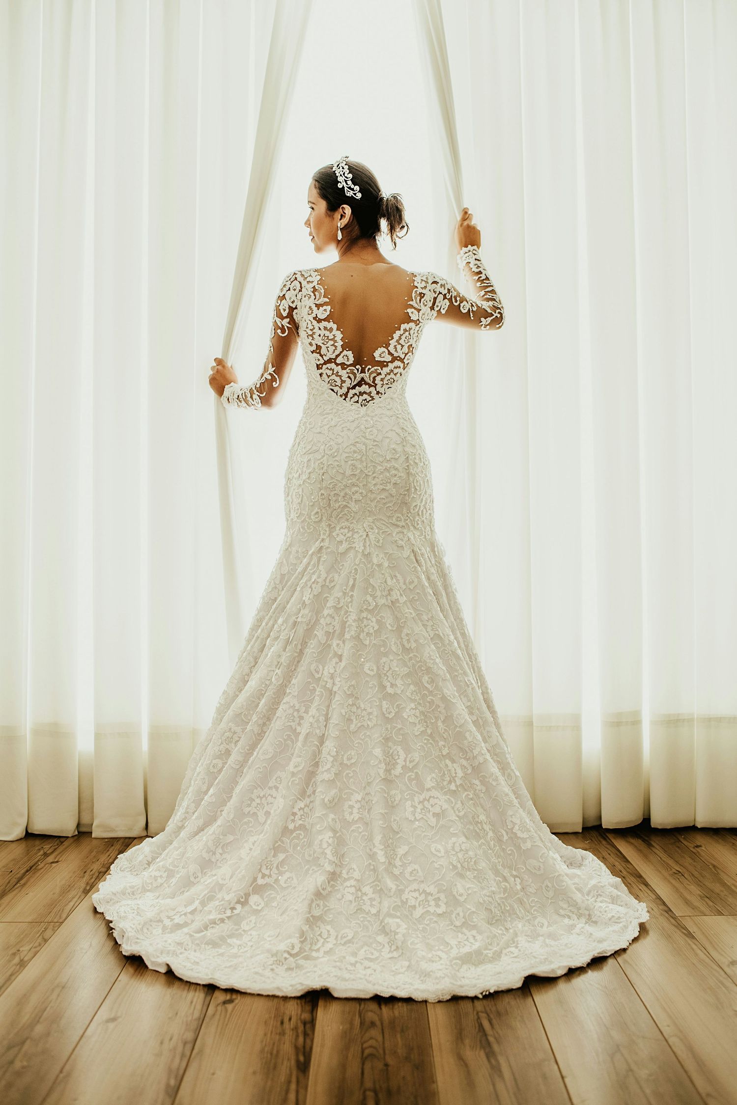 Bride in elegant long-sleeve lace mermaid wedding gown with deep V-back standing by sheer curtains.