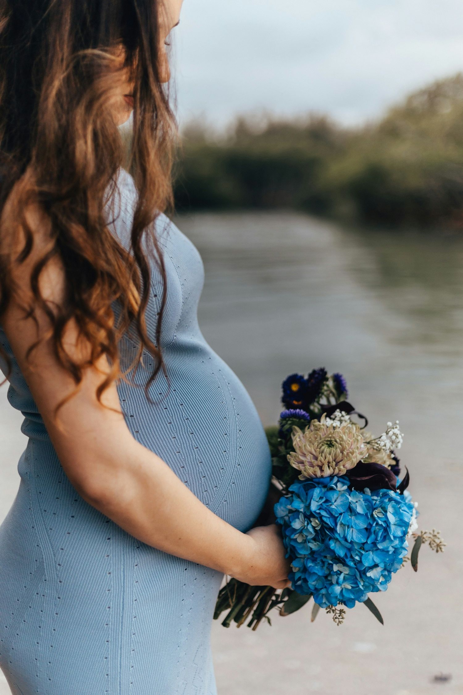 Pregnant woman in blue dress holding a bouquet of blue hydrangeas near a river outdoors.