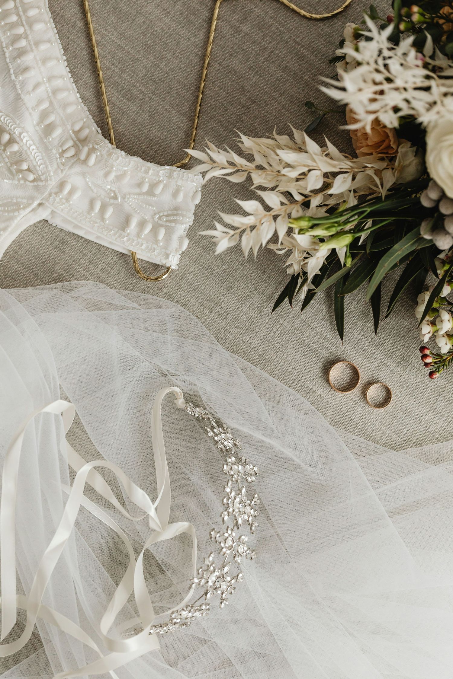 Bridal accessories flat lay featuring a crystal hair vine, veil, wedding rings, white dress, and floral bouquet.