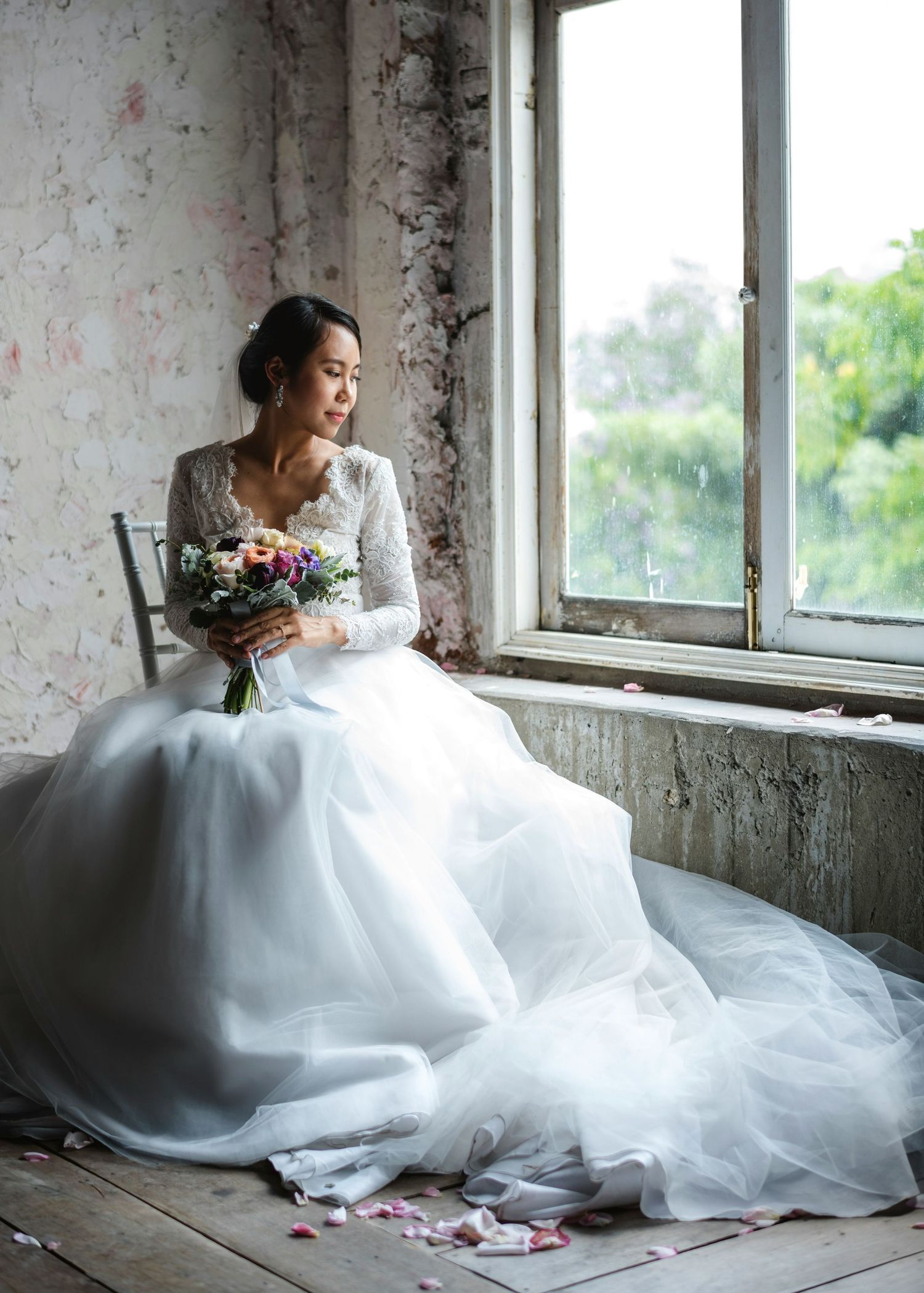 Bride in long-sleeve tulle gown holds colorful bouquet by rustic window.