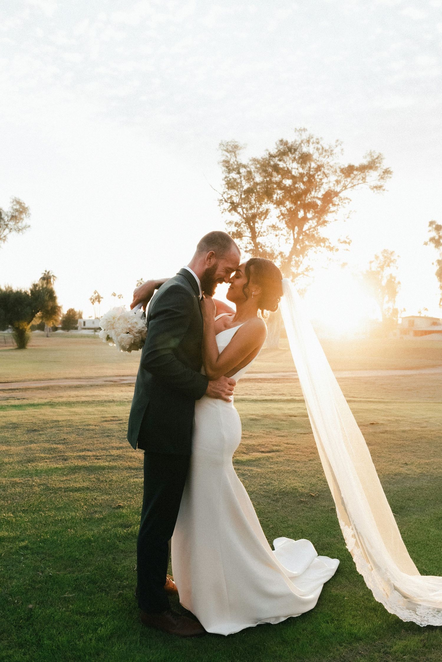 Bride and groom share a romantic forehead touch at golden hour on a lush green lawn, veil flowing in the breeze.