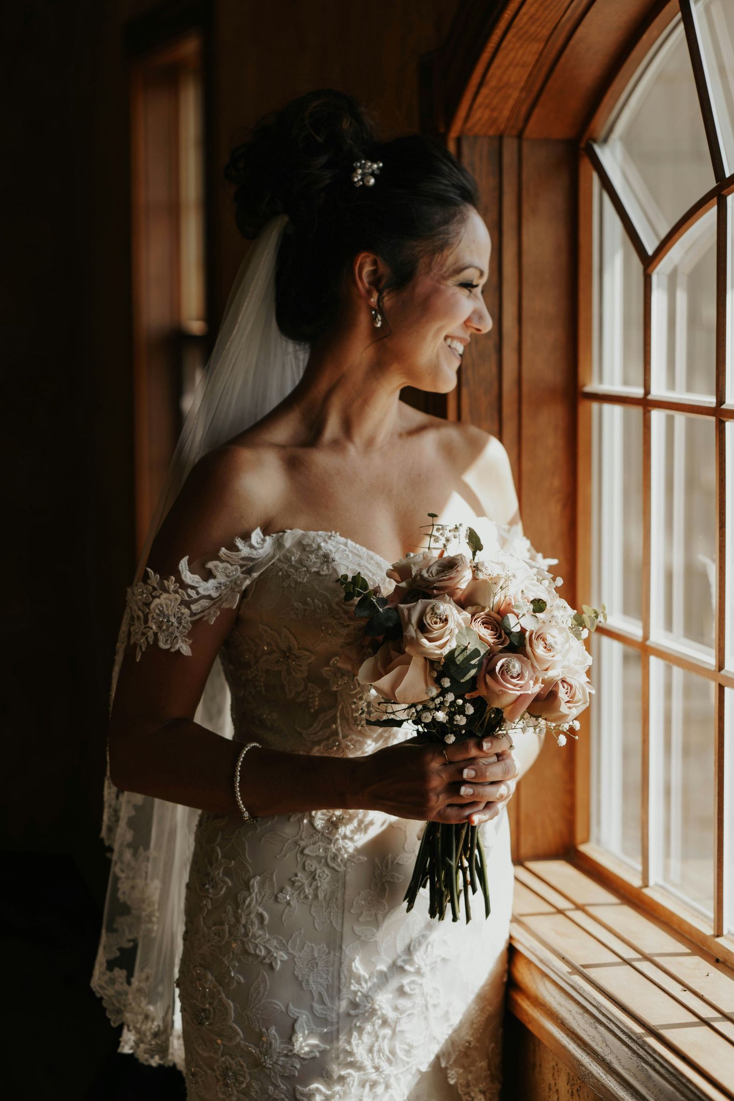 Elegant bride in off-shoulder lace gown holding pink rose bouquet by stained glass window.
