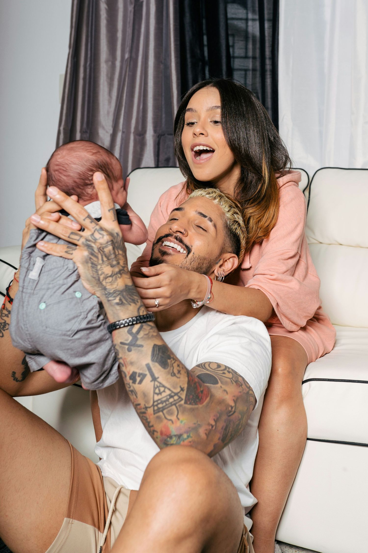 Tattooed father laughing while holding newborn baby with mother smiling behind him on living room floor.