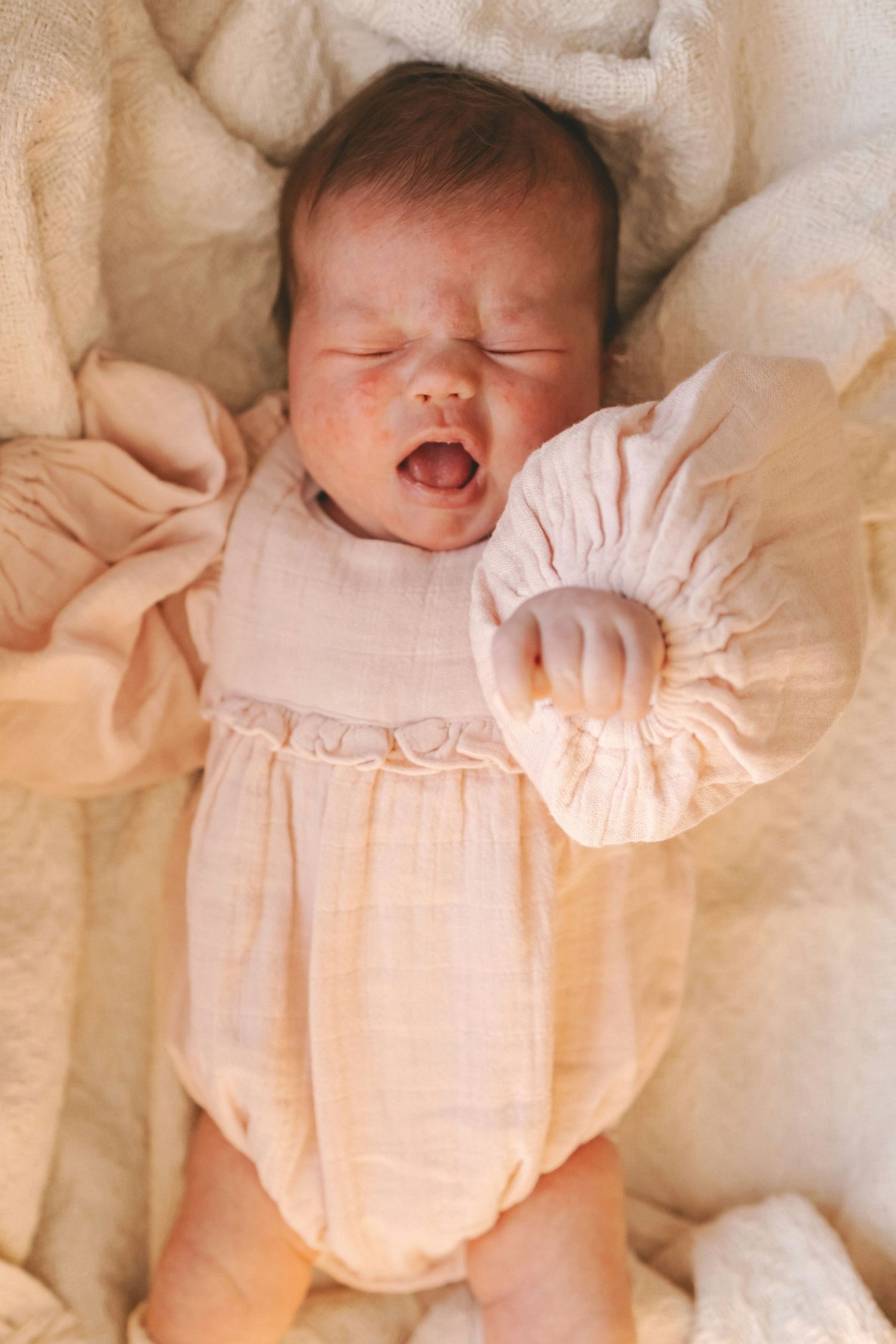 Yawning newborn baby in a pink muslin romper with ruffled sleeves lying on a cream blanket.