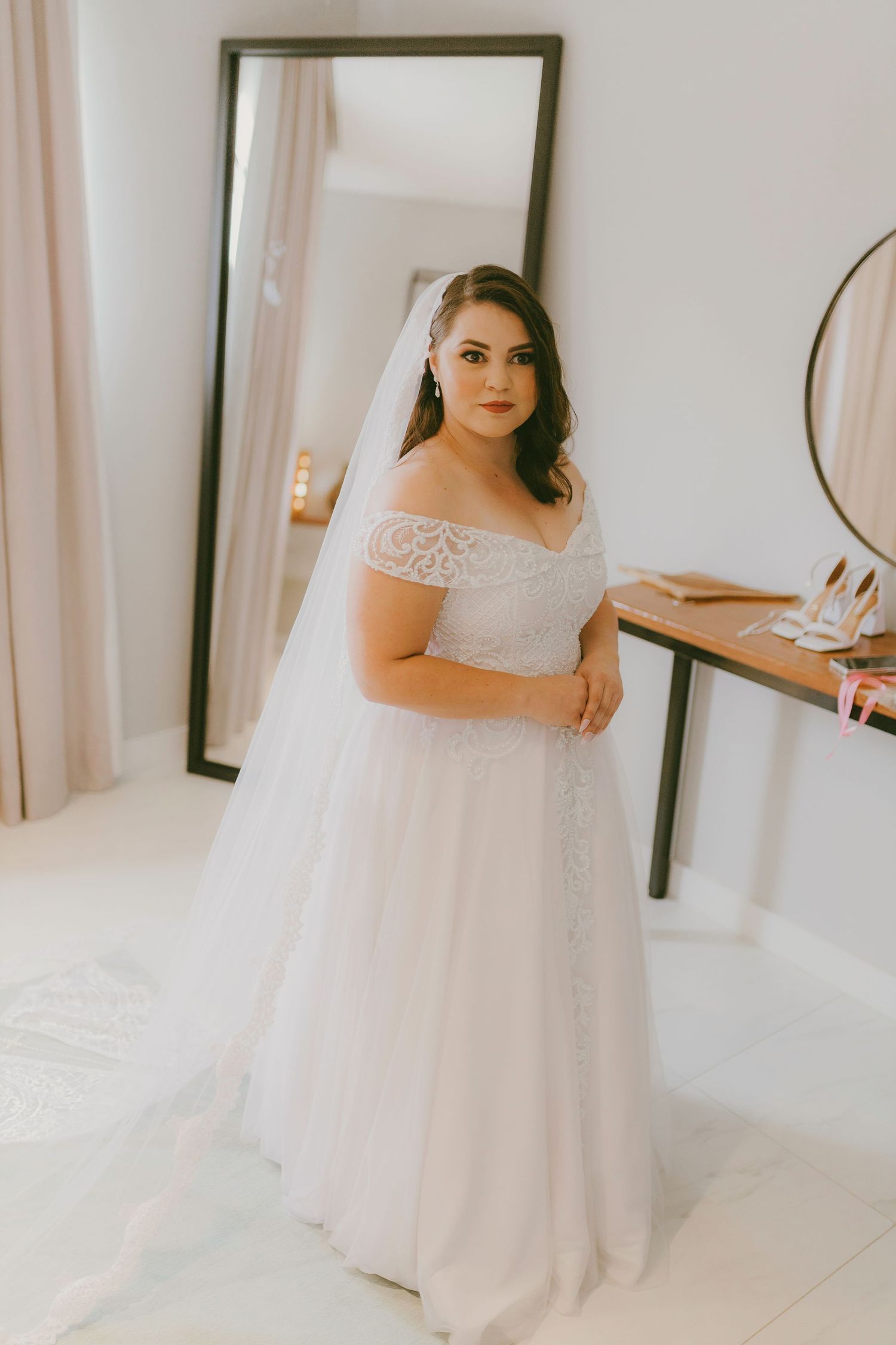 Bride in off-shoulder lace wedding gown with veil poses in bright dressing room before full-length mirror.