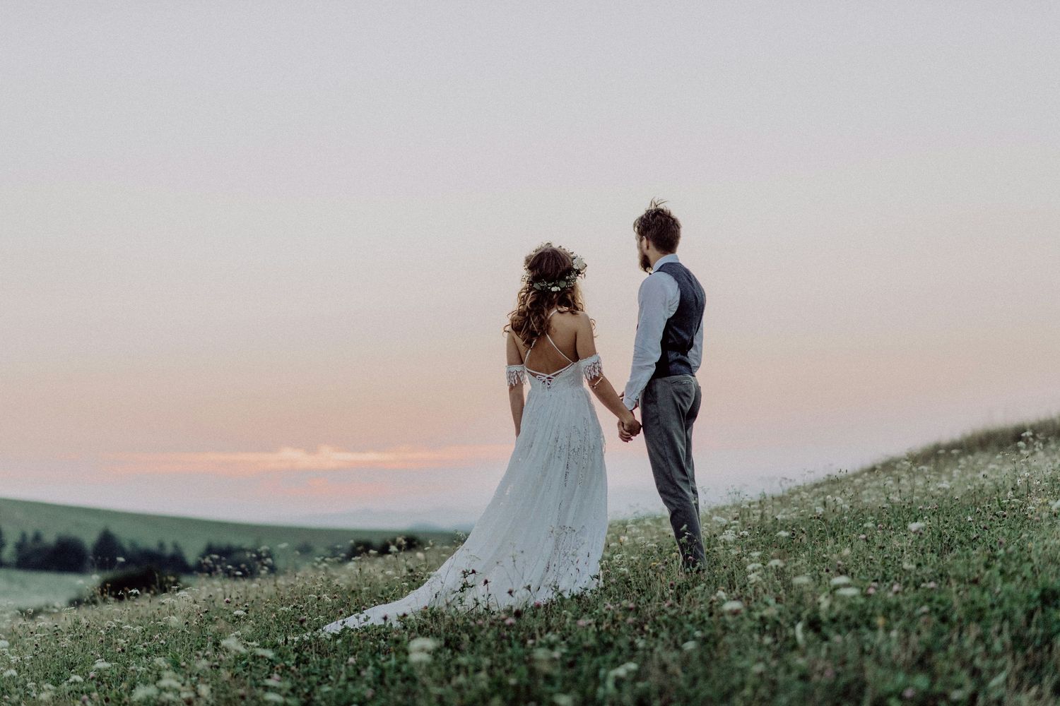 A bride and groom hold hands in a wildflower meadow at sunset during their outdoor wedding.