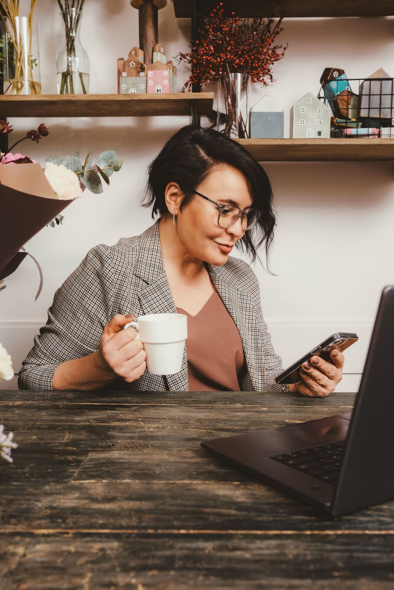 Stylish woman in blazer holding coffee mug and phone while working on laptop in cozy office.