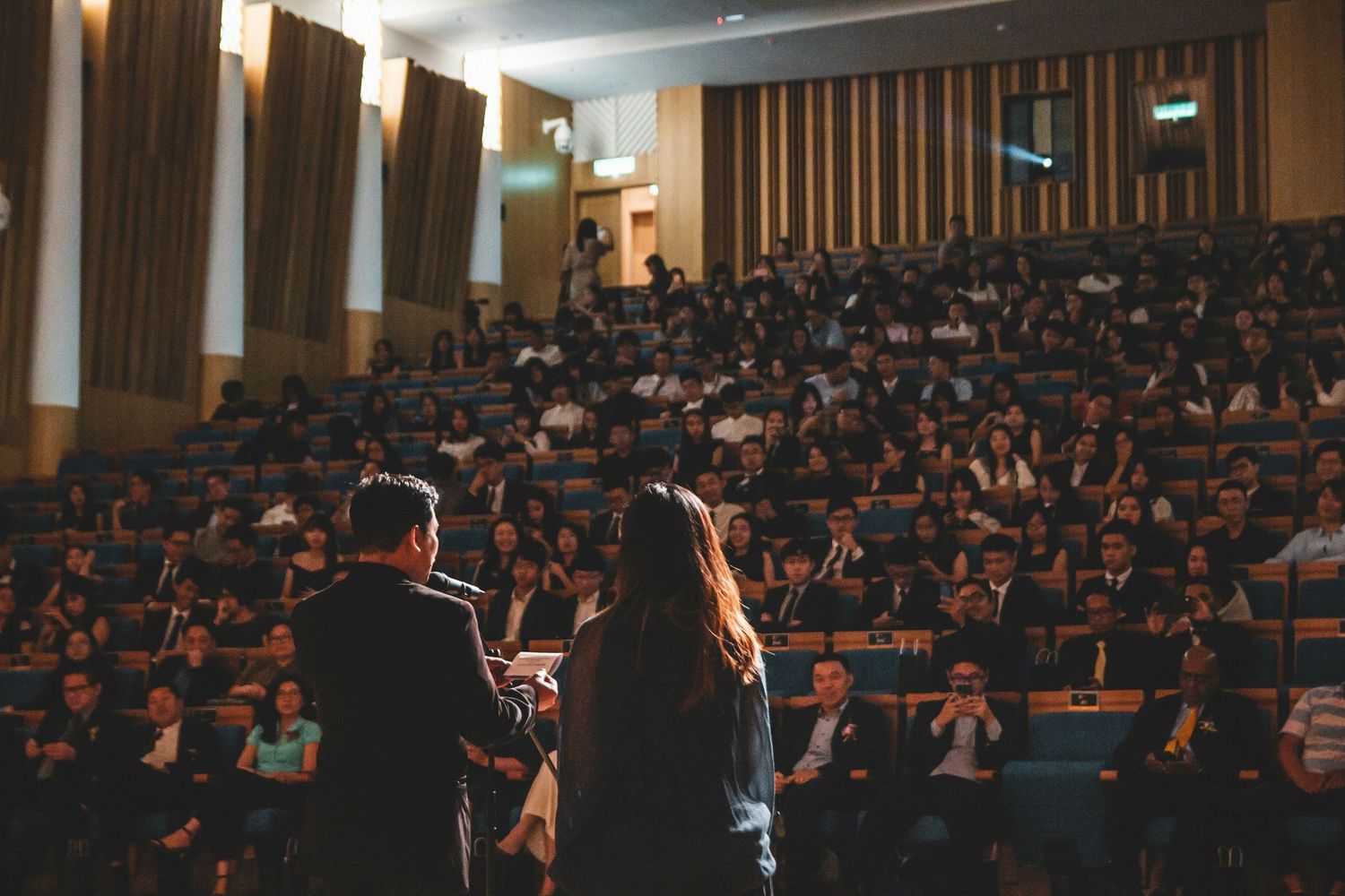 Two speakers present on stage to a large audience in a packed auditorium during a formal conference event.