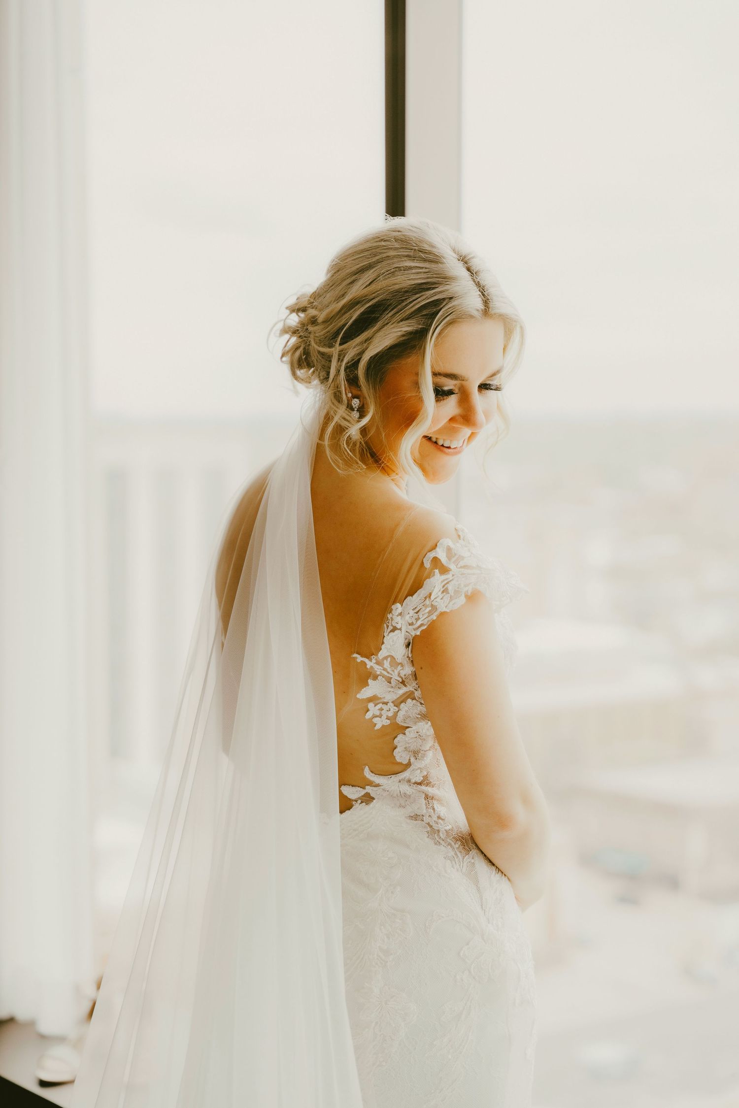 Smiling bride in backless lace wedding gown with flowing veil stands by window in soft natural light.