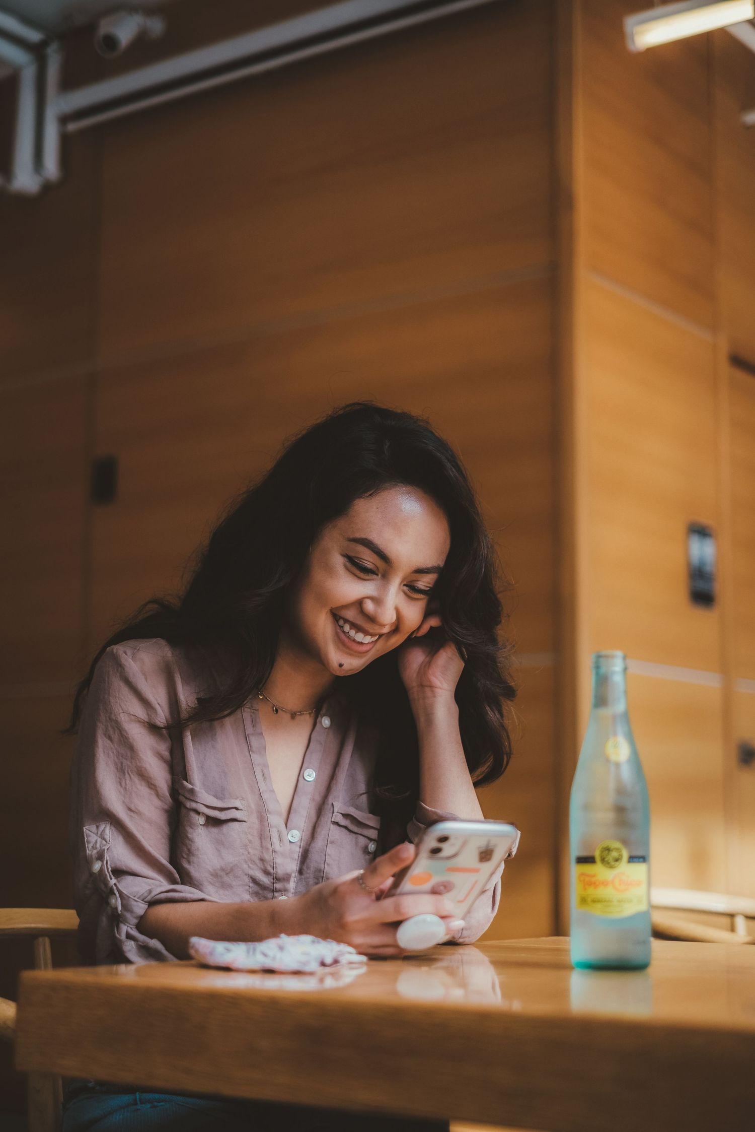Happy young woman laughing while using smartphone at wooden table with sparkling water bottle.