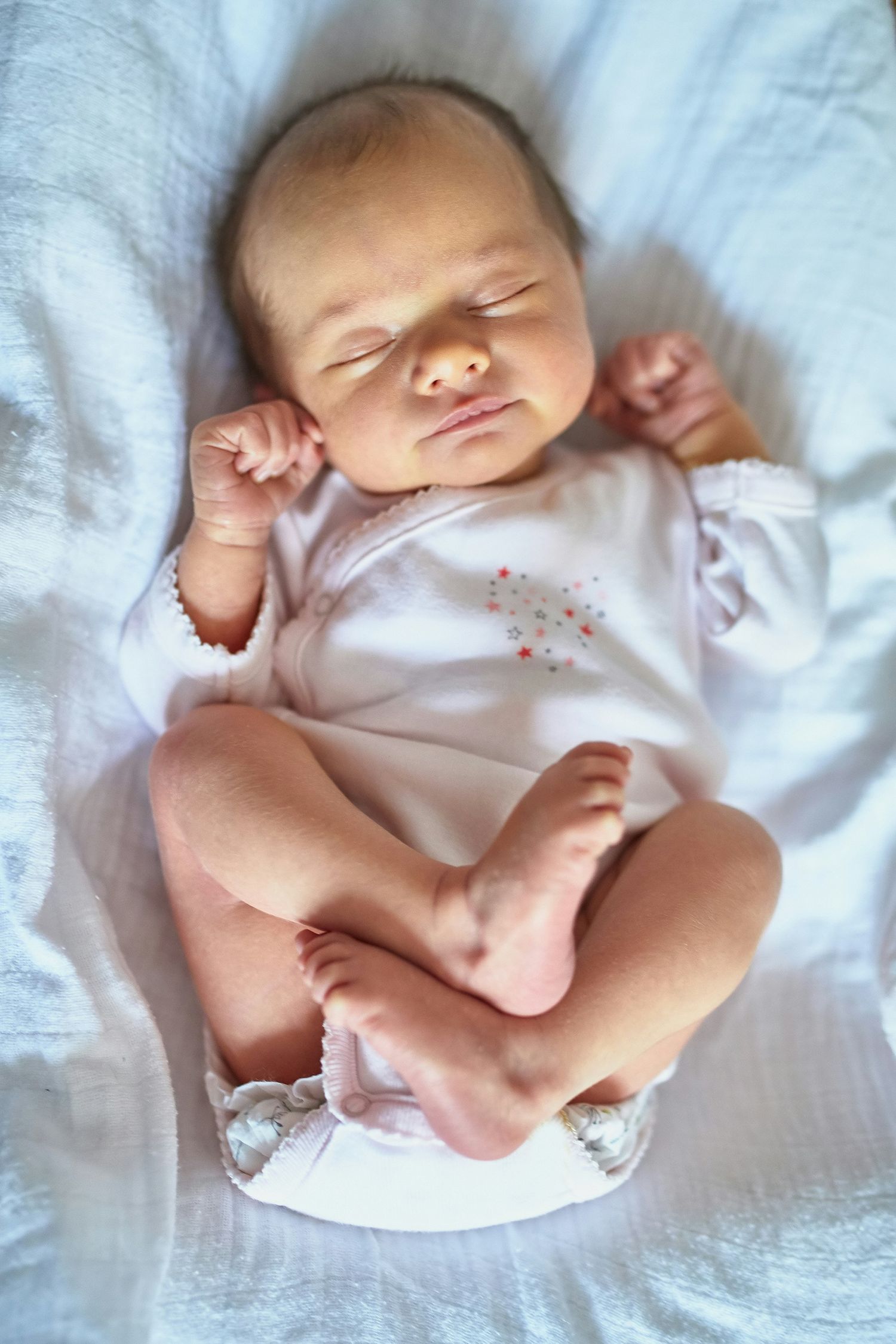 Sleeping newborn baby in a white onesie with tiny feet crossed lying on soft white blankets.