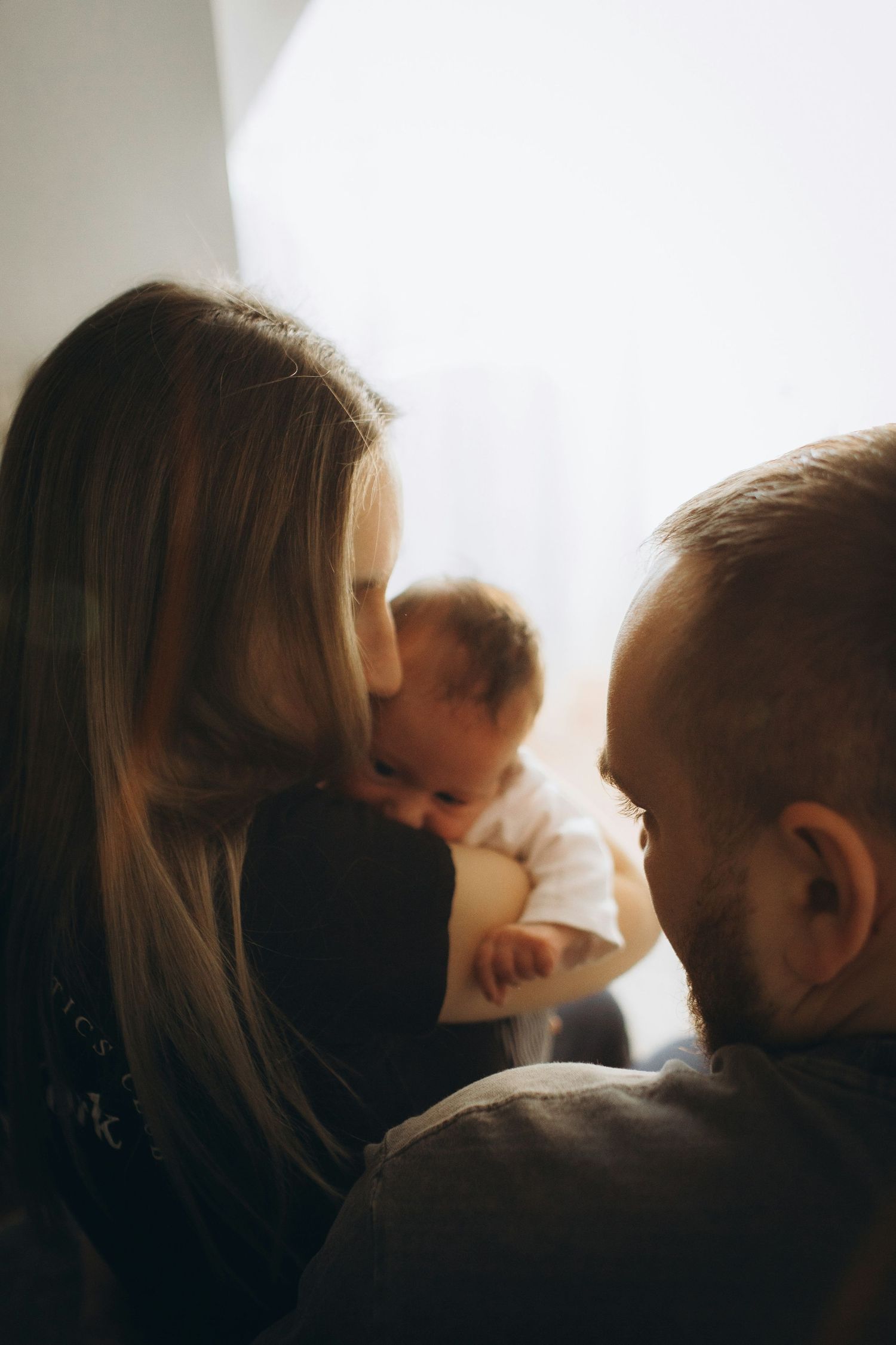 Mother kissing newborn baby while father looks on lovingly near bright window.