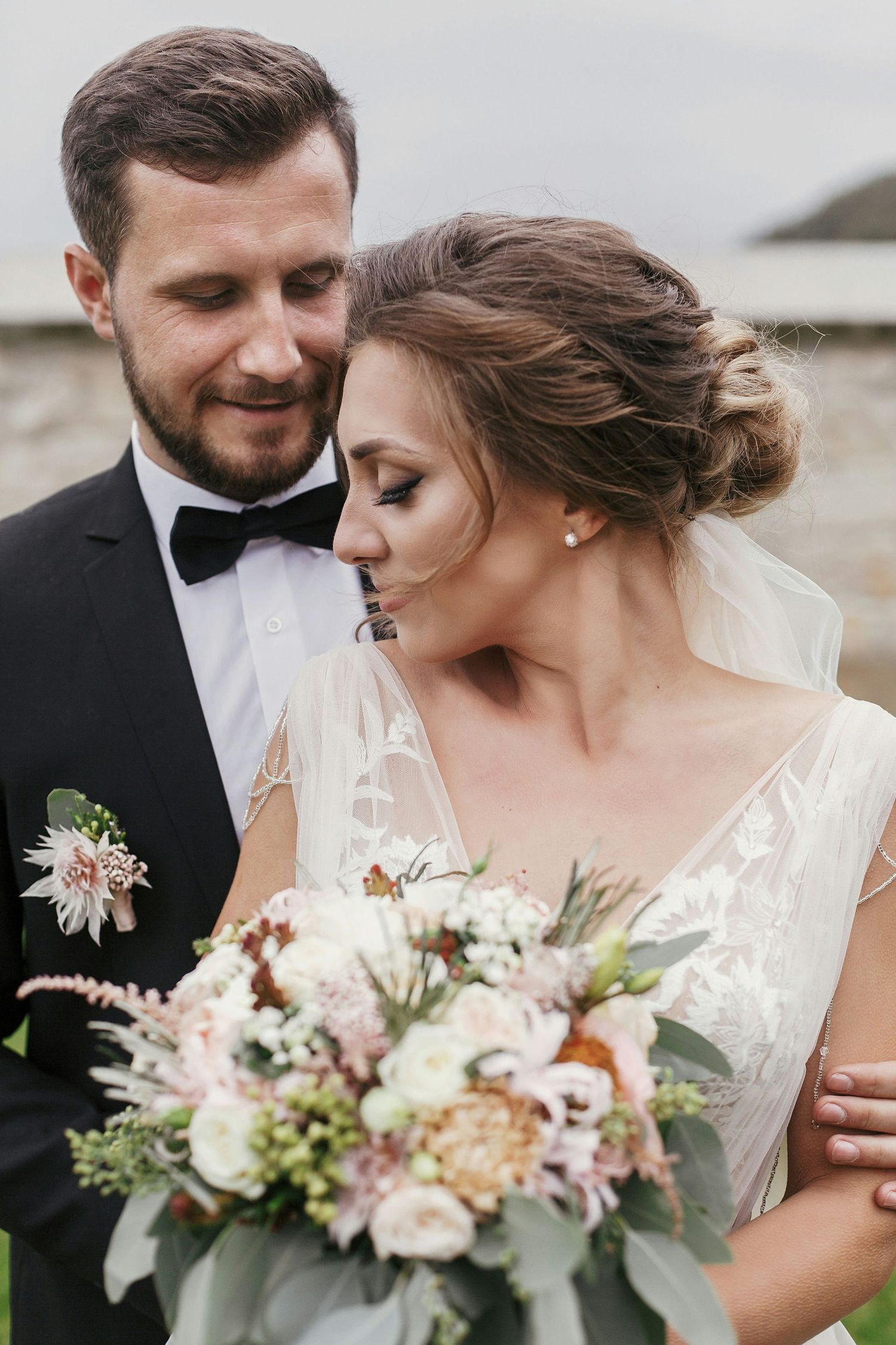Groom in black tuxedo embracing bride in lace gown holding soft pink and white floral bouquet outdoors.