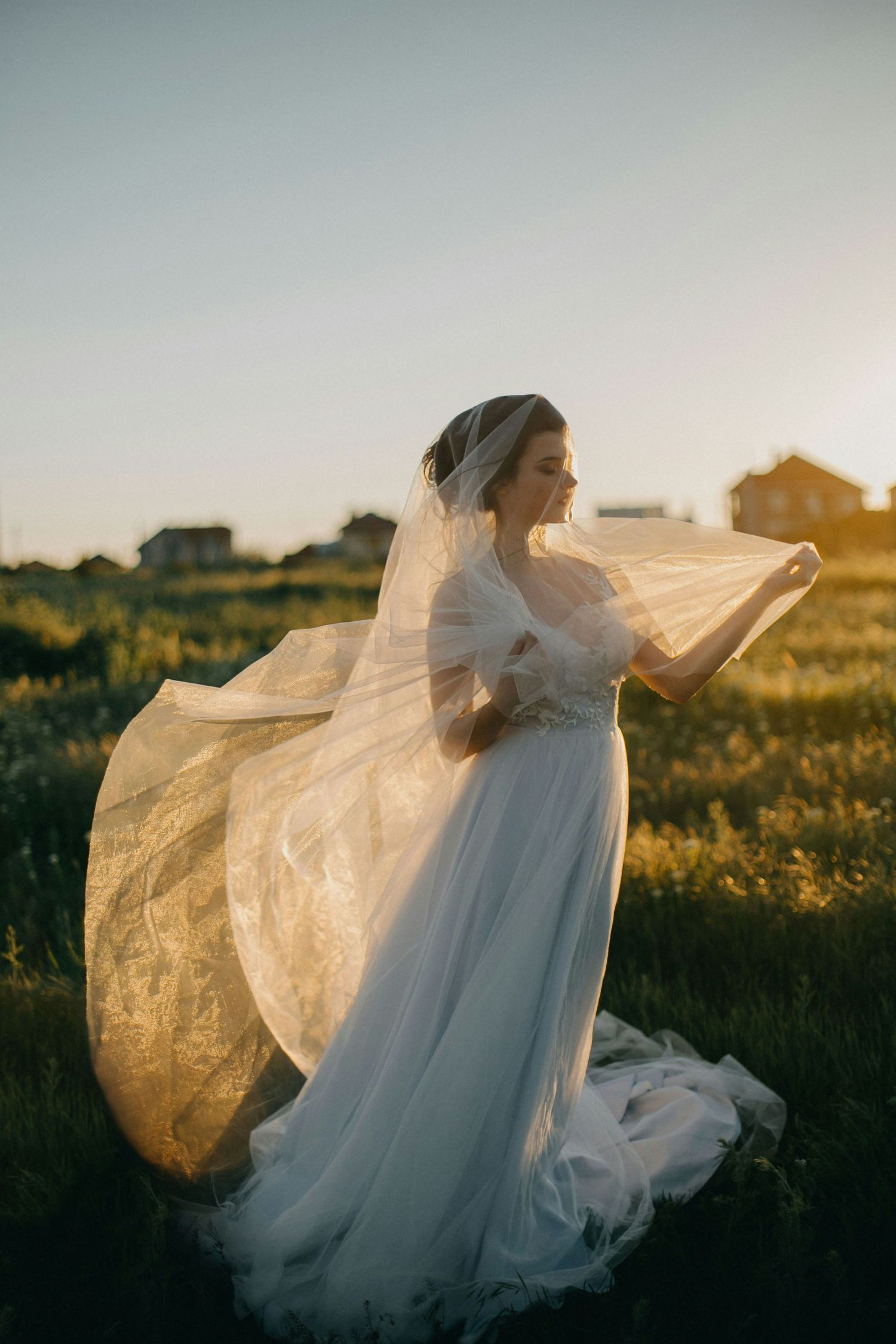 Bride in flowing white lace wedding dress holding veil in golden sunset field.