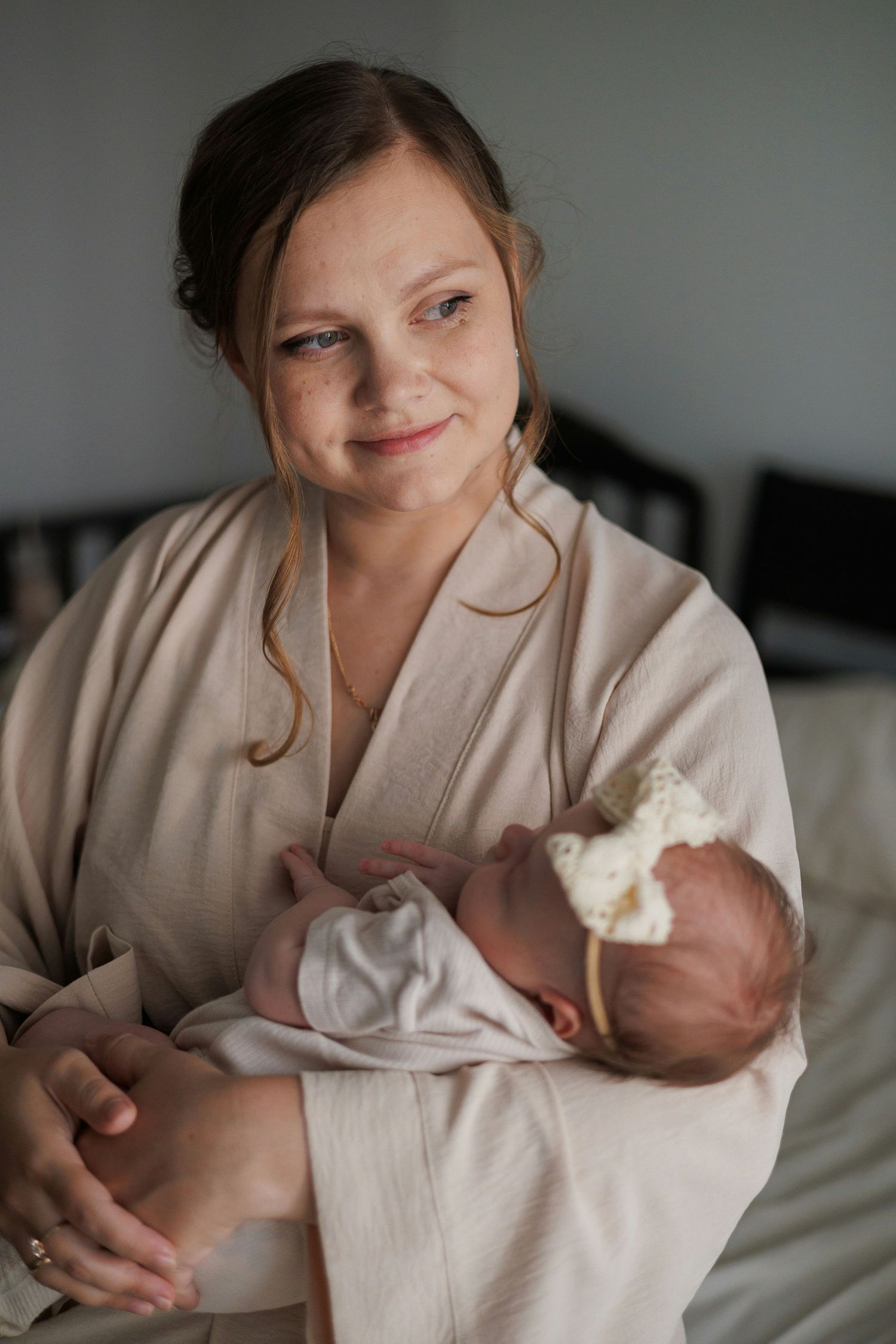Young mother in beige robe smiling while holding sleeping newborn with bow headband.