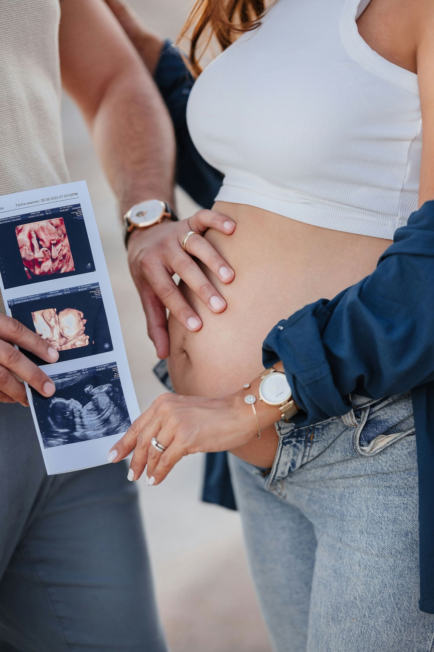 Couple holding ultrasound images while touching pregnant belly outdoors.