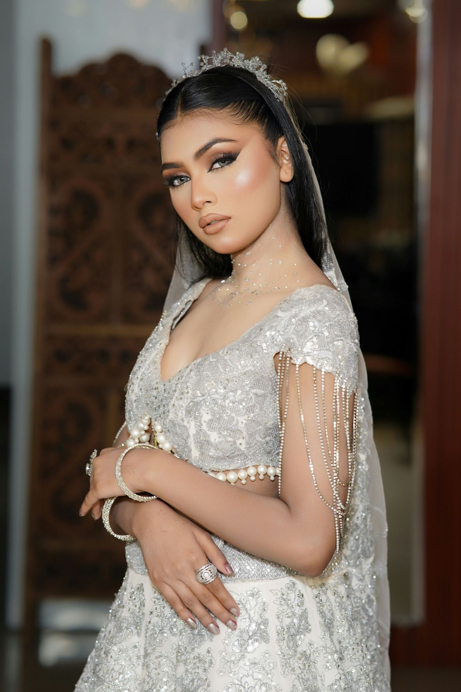 Glamorous bride in silver embroidered gown with pearl accessories and crystal crown indoors.