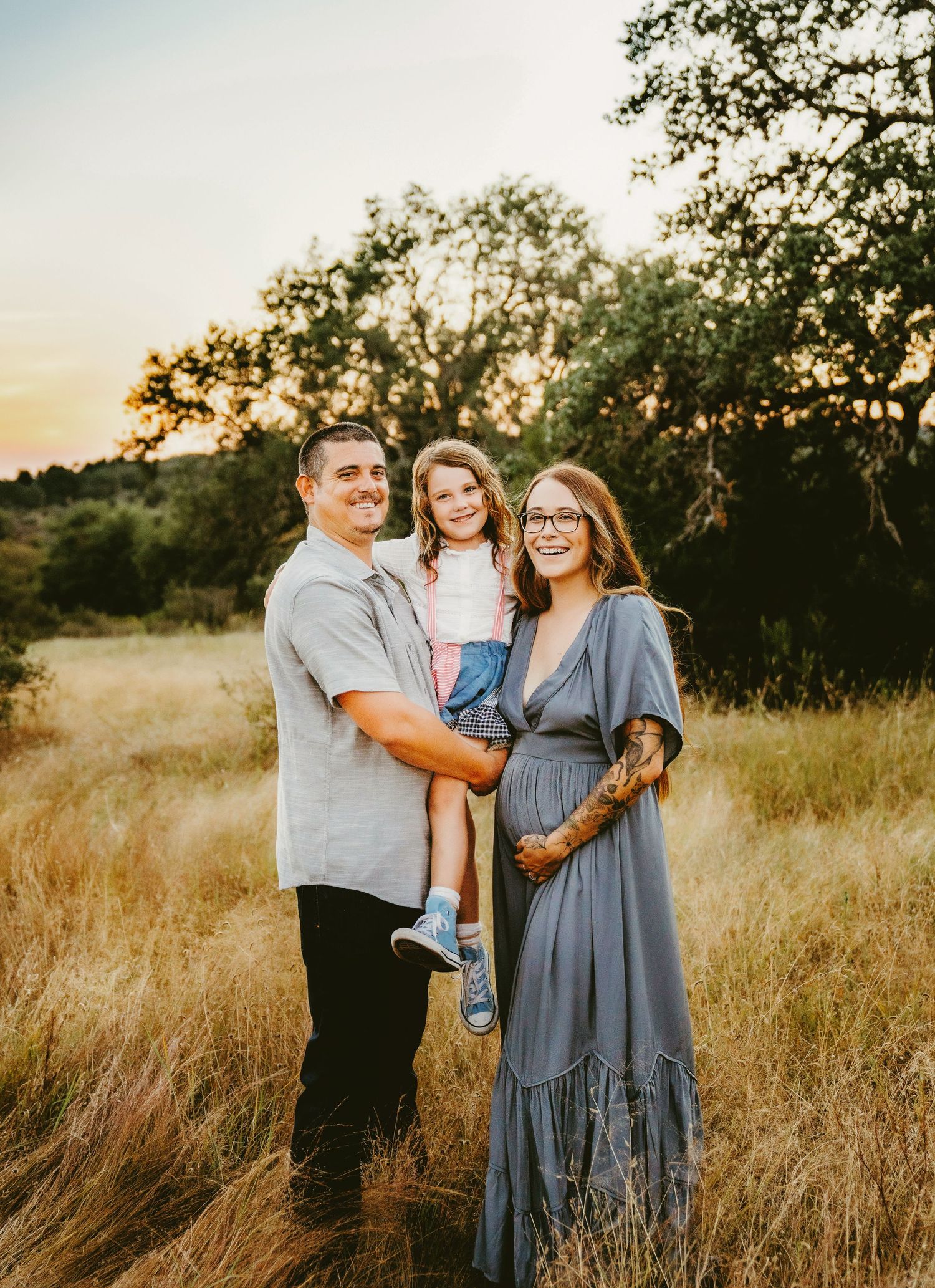 A happy family of three poses in a golden field at sunset, with a man holding a young girl and a pregnant woman in a blue dress.
