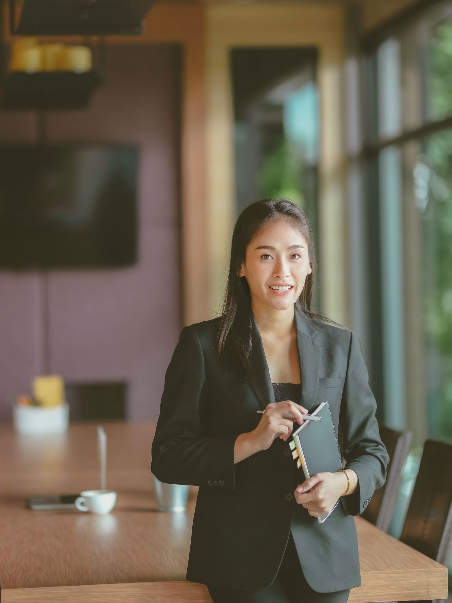 Professional businesswoman in black suit holding notebook and pen, smiling confidently in modern office boardroom.