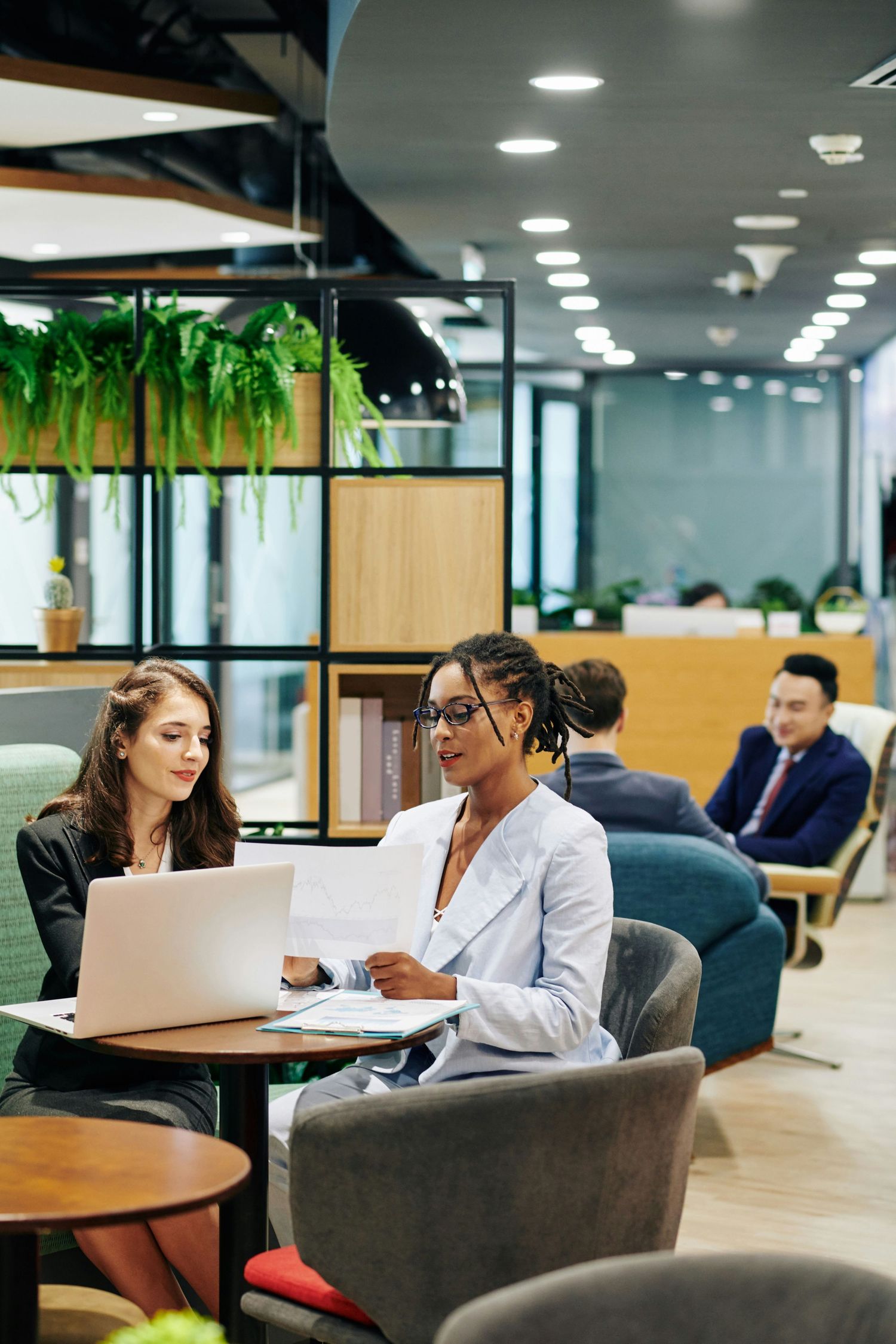 Two professional women collaborate at a laptop in a modern office lounge, with colleagues working in the background.