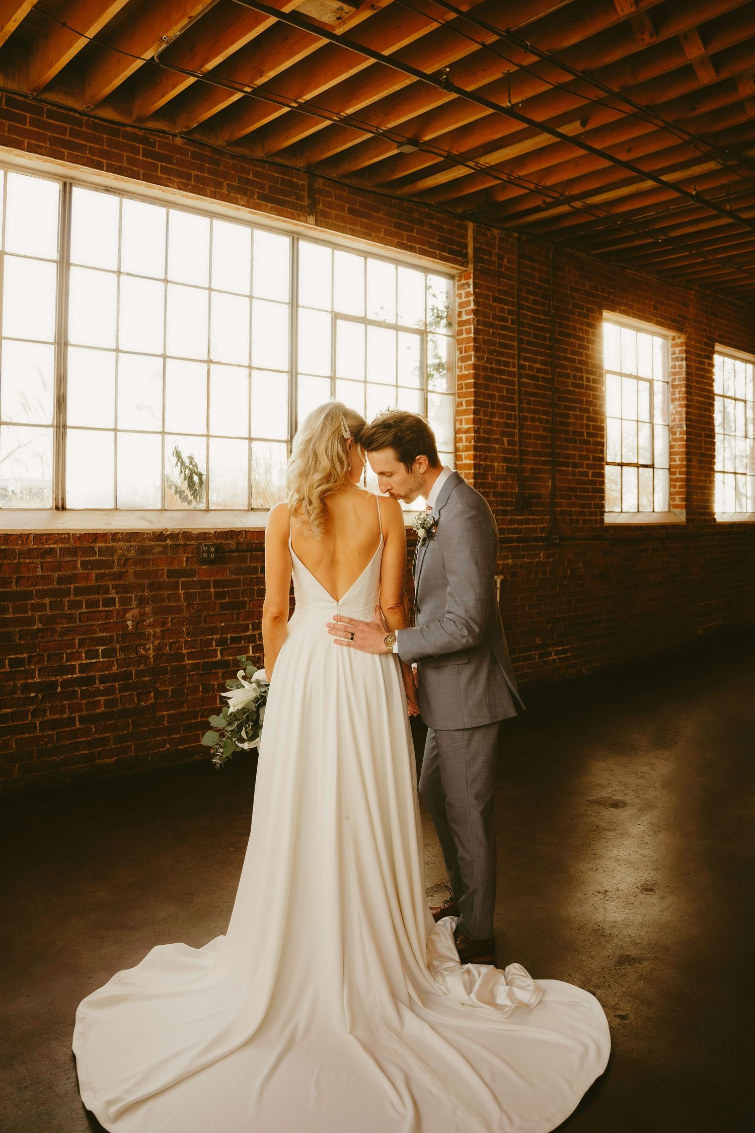 Bride in backless white gown and groom in gray suit share intimate moment in rustic brick venue with large windows.