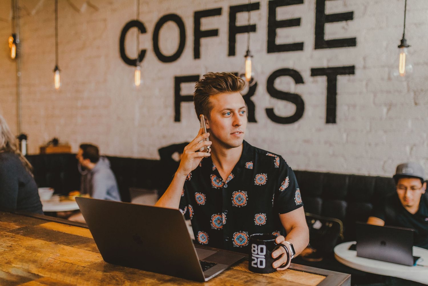 Young man talking on phone at laptop in trendy coffee shop with Coffee First wall sign.